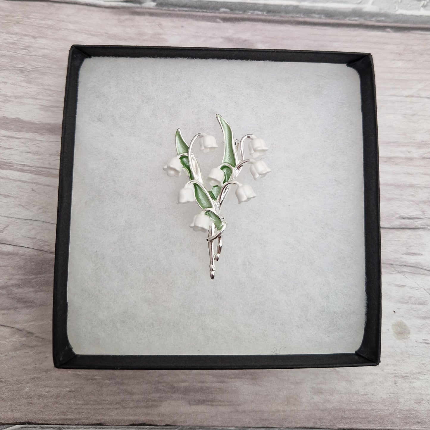 Photo of a spray of Lily of the Valley white flowers and green leaves formed as a ladies brooch with a silver stem in a gift box.
