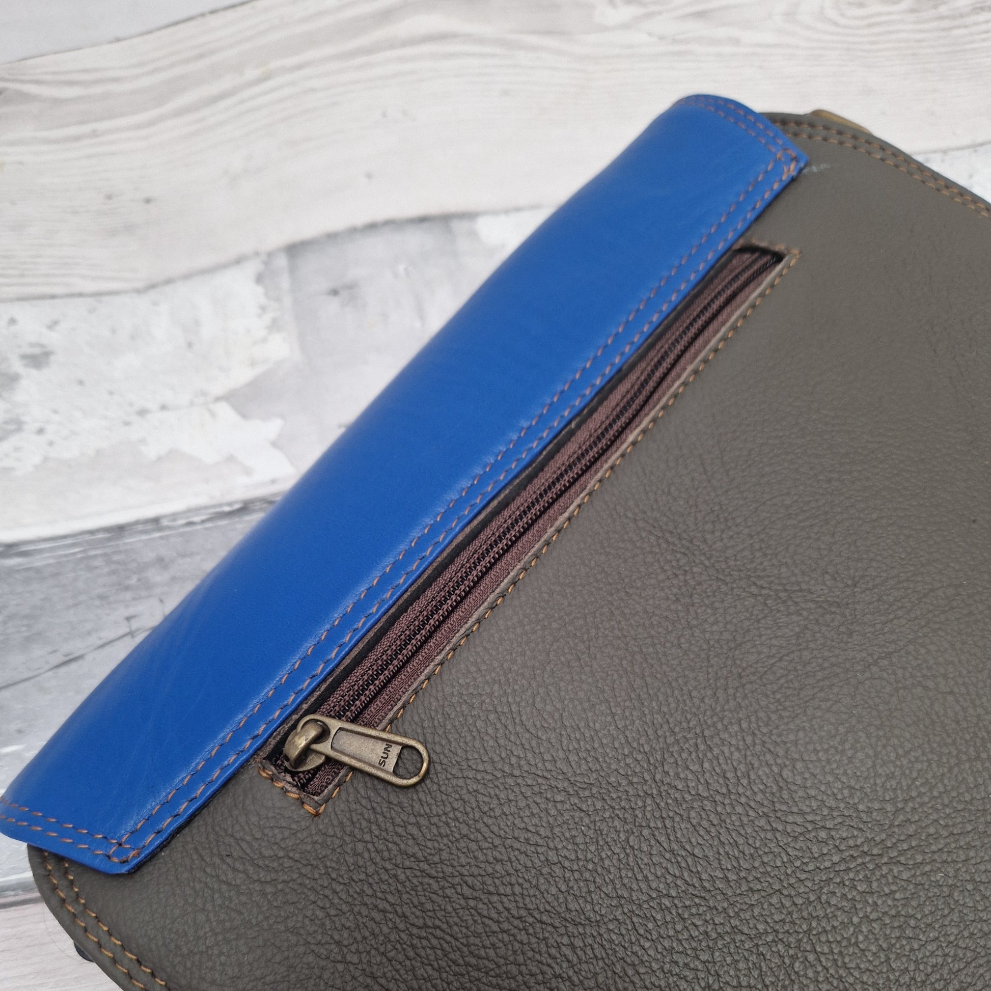 Gray leather bag with a blue flap and zipper on a light wooden surface