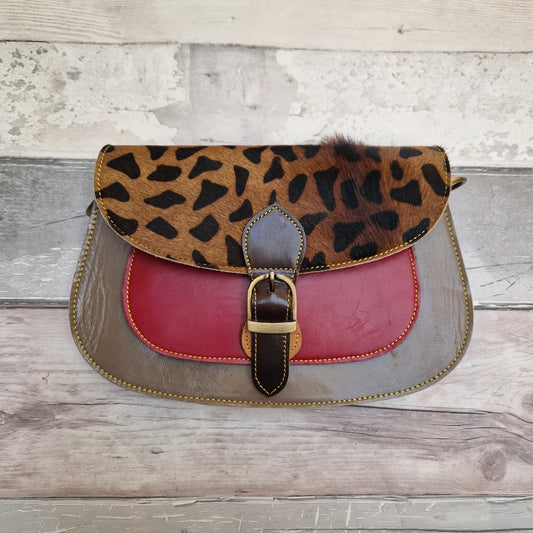 A vintage-style saddle handbag with a giraffe-print flap, a red leather center panel, and a gray leather base, featuring a brown leather strap and gold-tone buckle, displayed on a rustic wooden plank background.