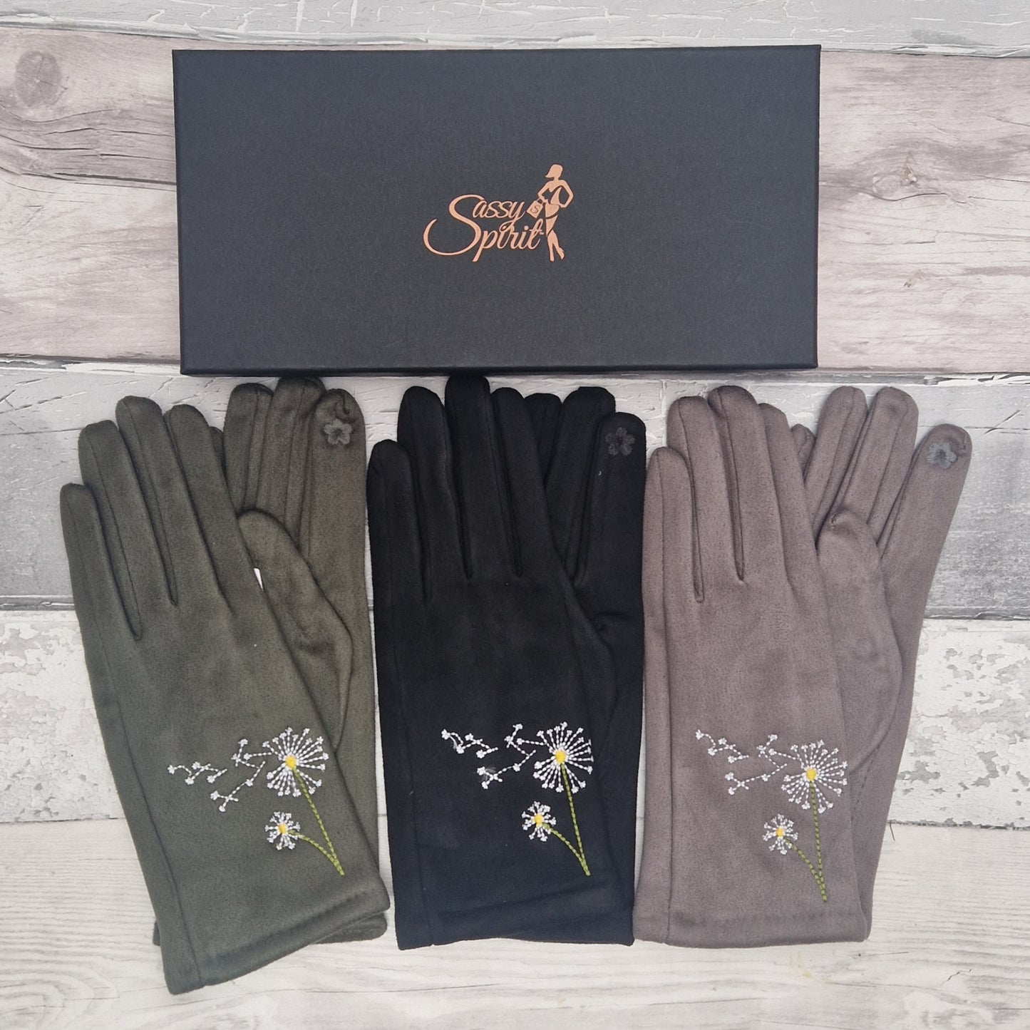 Three pairs of gloves with dandelion designs on a wooden surface, branded 'Sassy Spirit'.