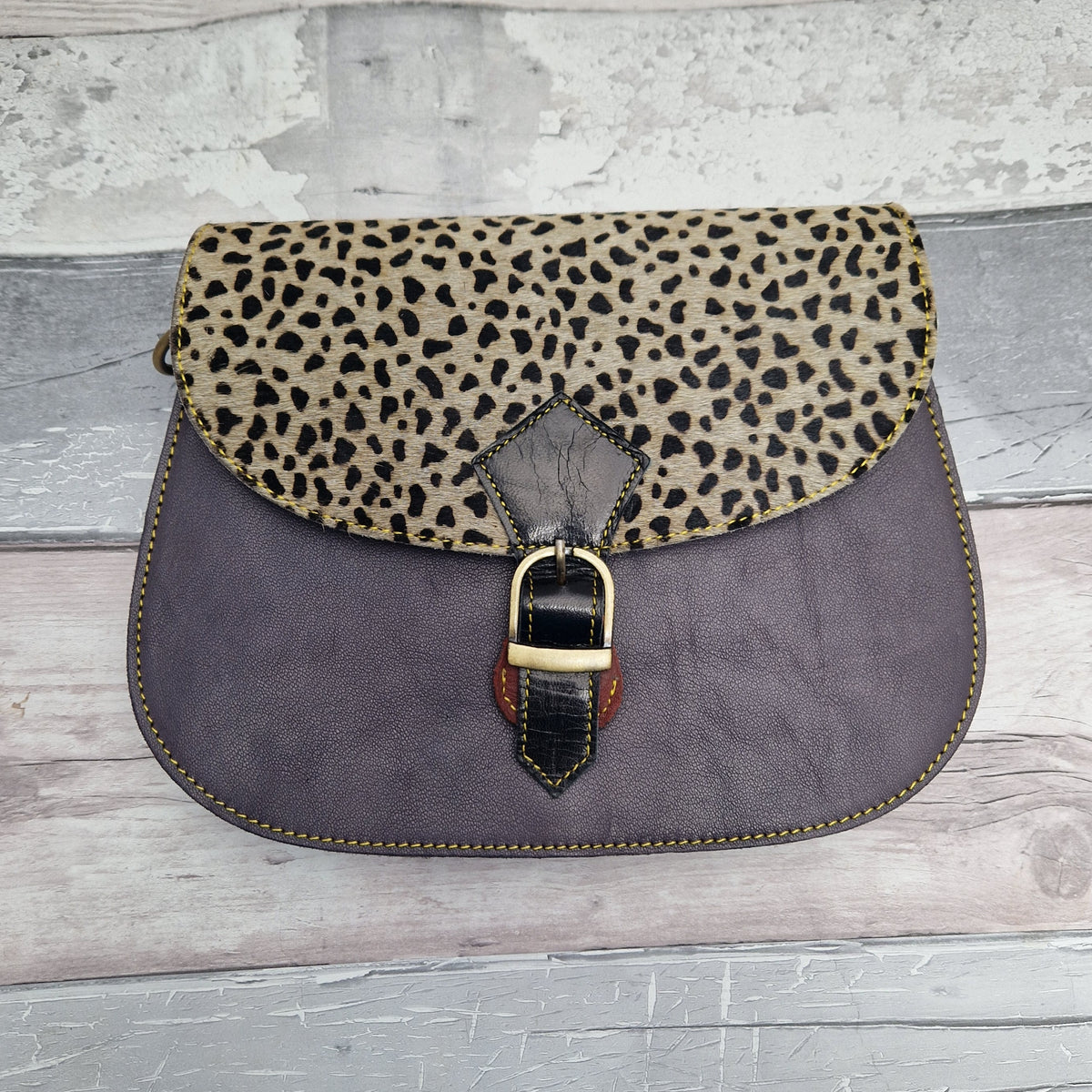 A vintage-style saddle bag featuring a dark gray leather body, a leopard-print flap, and a gold-tone buckle closure, displayed on a rustic gray wood plank surface.