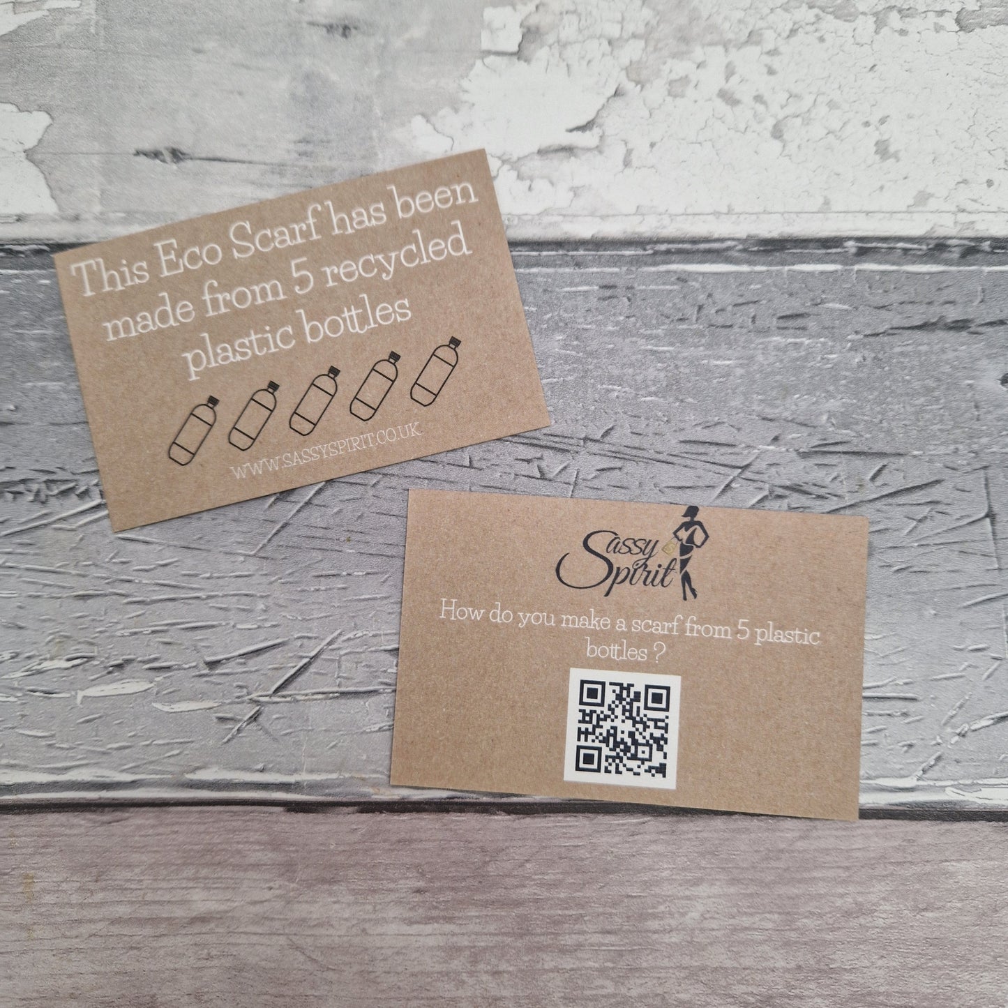 Two brown cards on a textured gray surface, one with text about recycled plastic bottles and the other with a QR code and Sassy Spirit logo.
