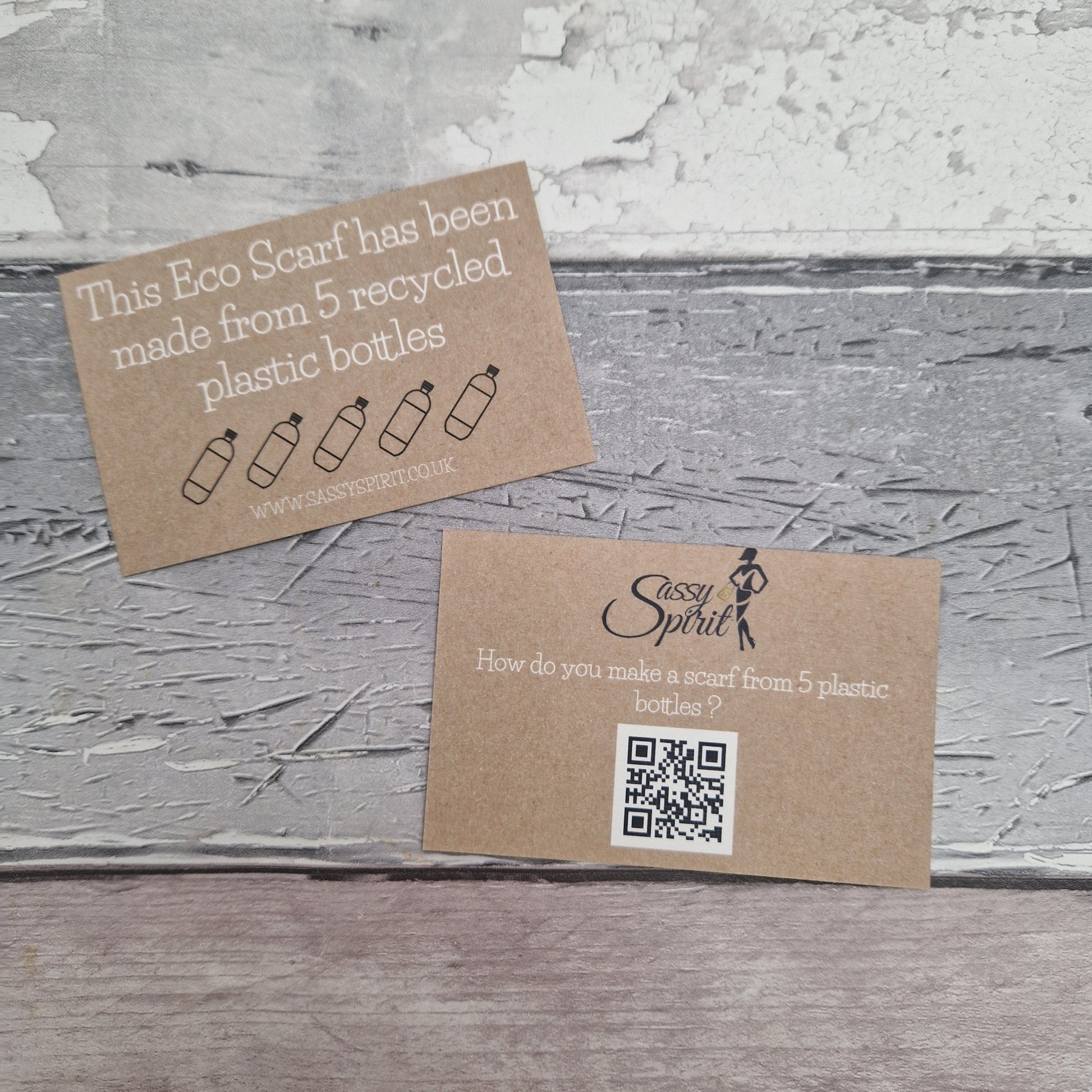Two brown cards on a textured gray surface, one with text about recycled plastic bottles and the other with a QR code and Sassy Spirit logo.