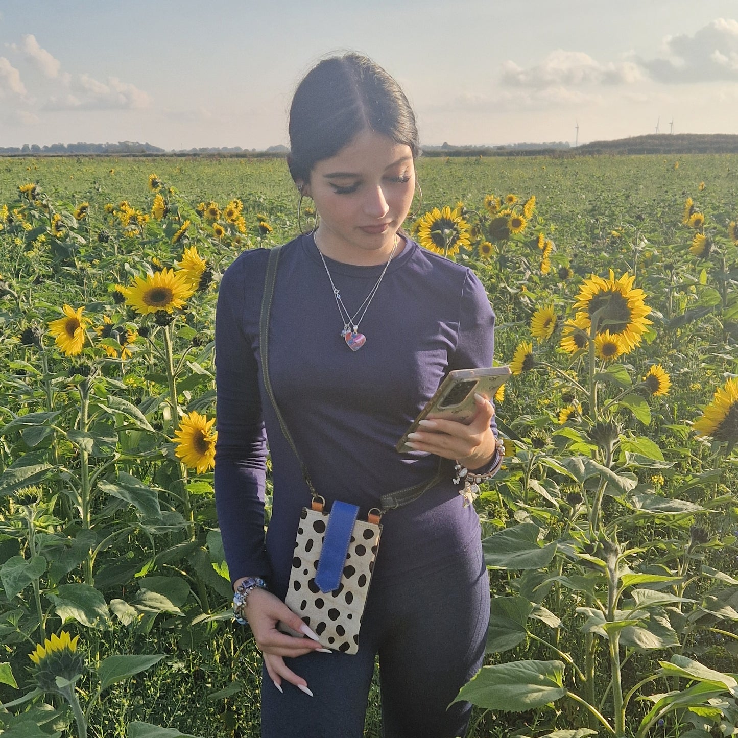 Woman standing in a sunflower field using a phone wearing a crossbody spotted pouch.