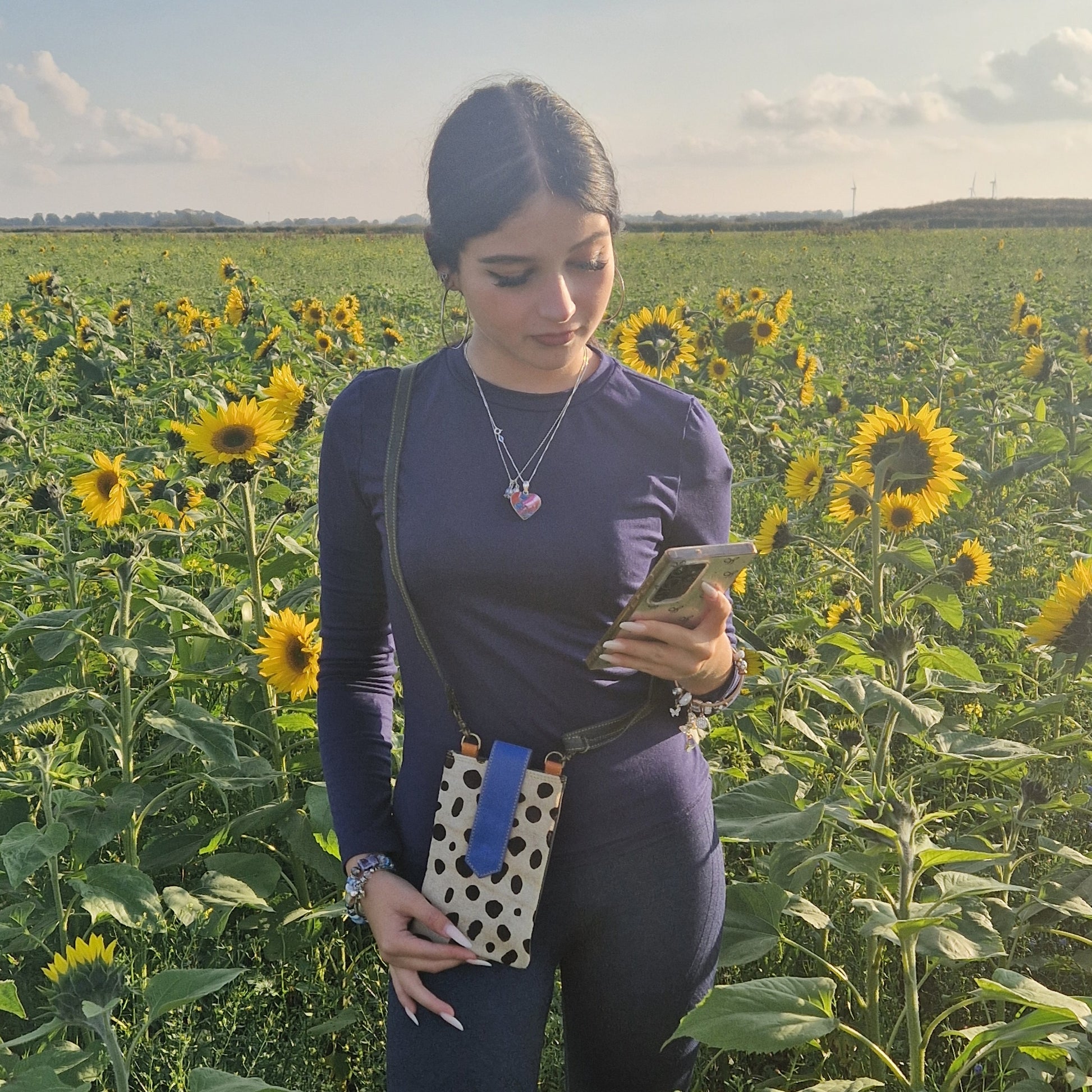 Woman standing in a sunflower field using a phone wearing a crossbody spotted pouch.
