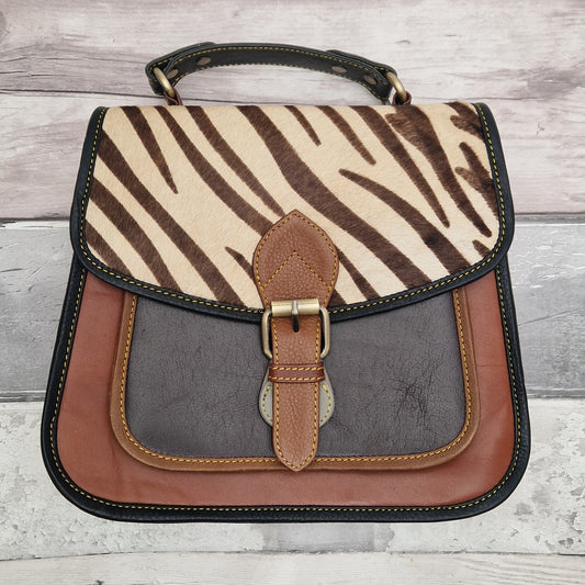 Brown saddlebag made up of multi-tonal sections and finished with a zebra print panel. Buckle front and top carry handle.