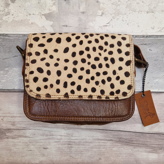 A small shoulder bag with a cream and black spotted pattern on the flap and dark brown textured leather on the front and bottom, shown on a rustic wood background with a hanging brand tag.