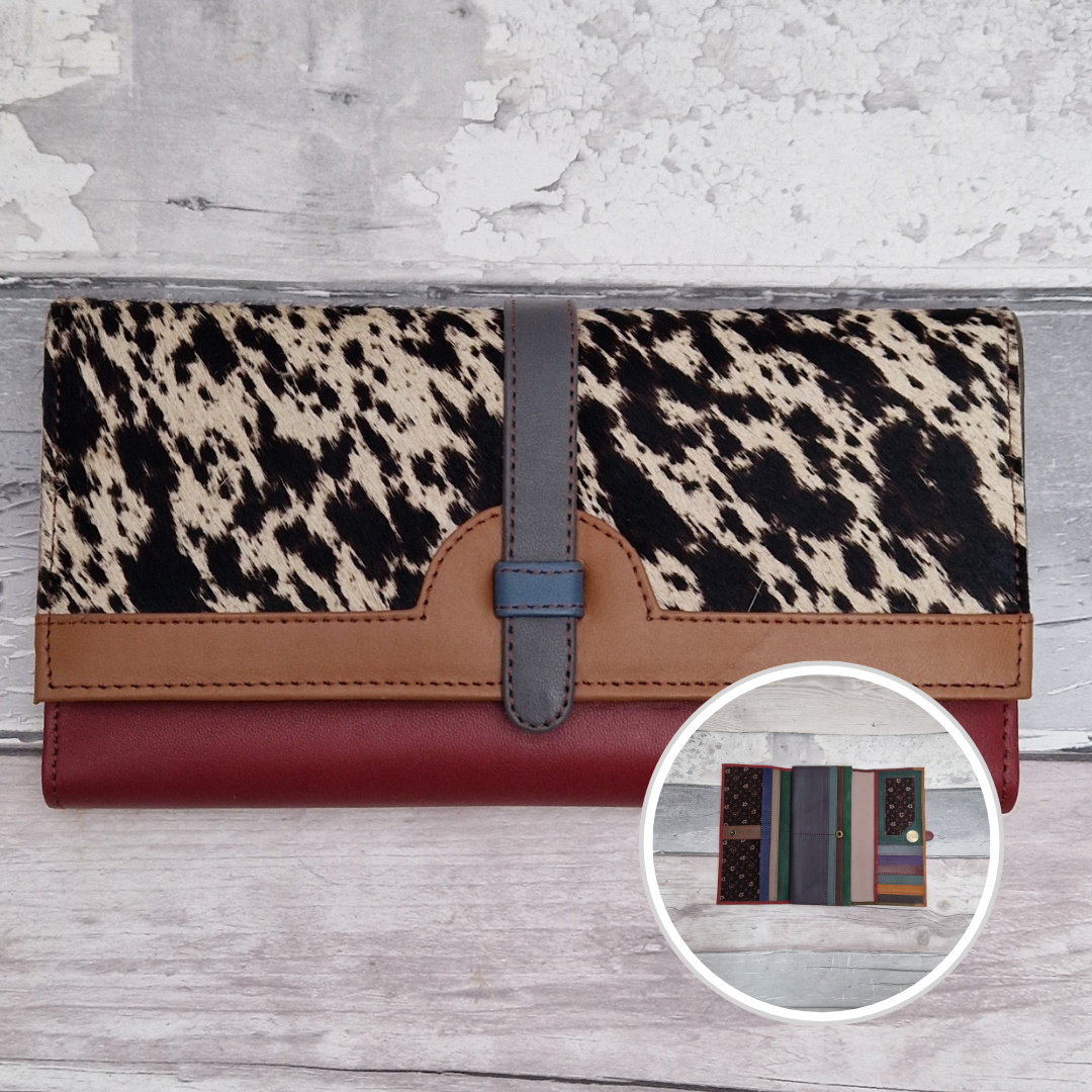 A long wallet with a black and beige animal print top flap, tan leather trim, a gray vertical strap, and a burgundy red lower section, shown on a light wood background with a circular inset showing the interior compartments.