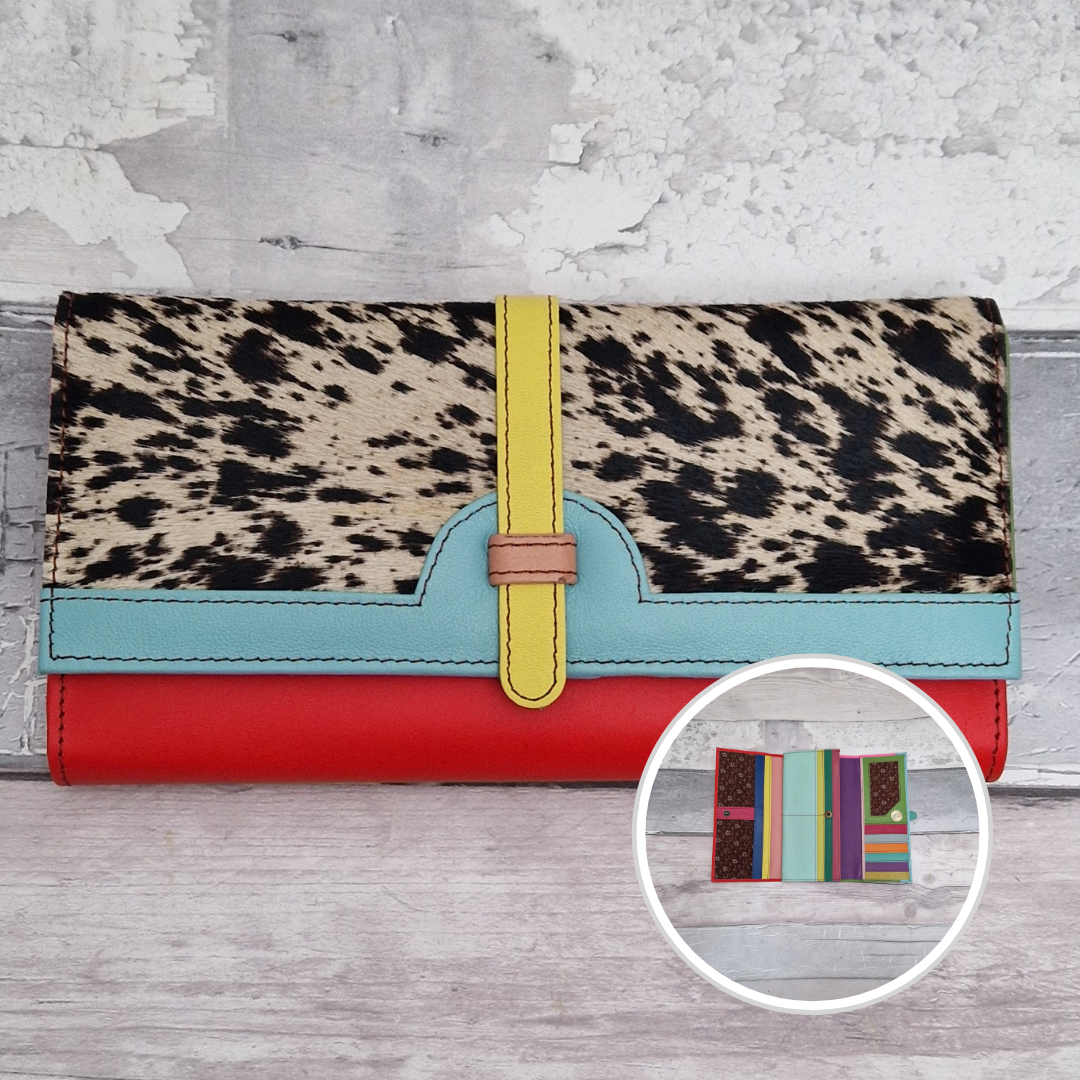 A multi-colored clutch wallet featuring a cow-print top flap with a yellow strap closure, a light blue accent band, and a red base, displayed on a light gray wood background with a circular inset showing its interior compartments.
