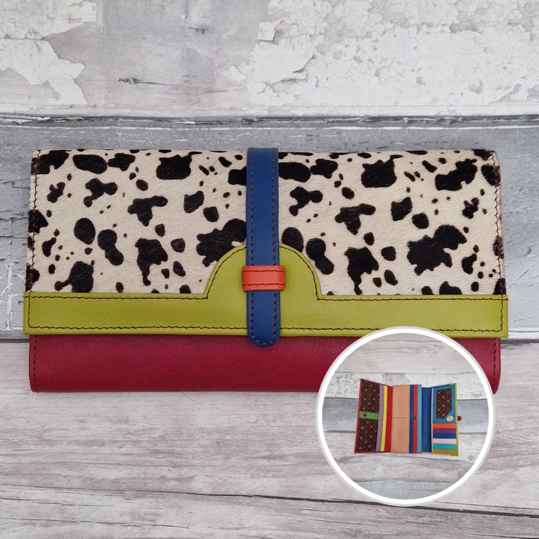 A multi-colored wallet featuring a cow-print top half, a lime green accent band, a red bottom half, and a blue strap closure with an orange accent. Shown on a light gray wood background with a circular inset showing the interior card slots.
