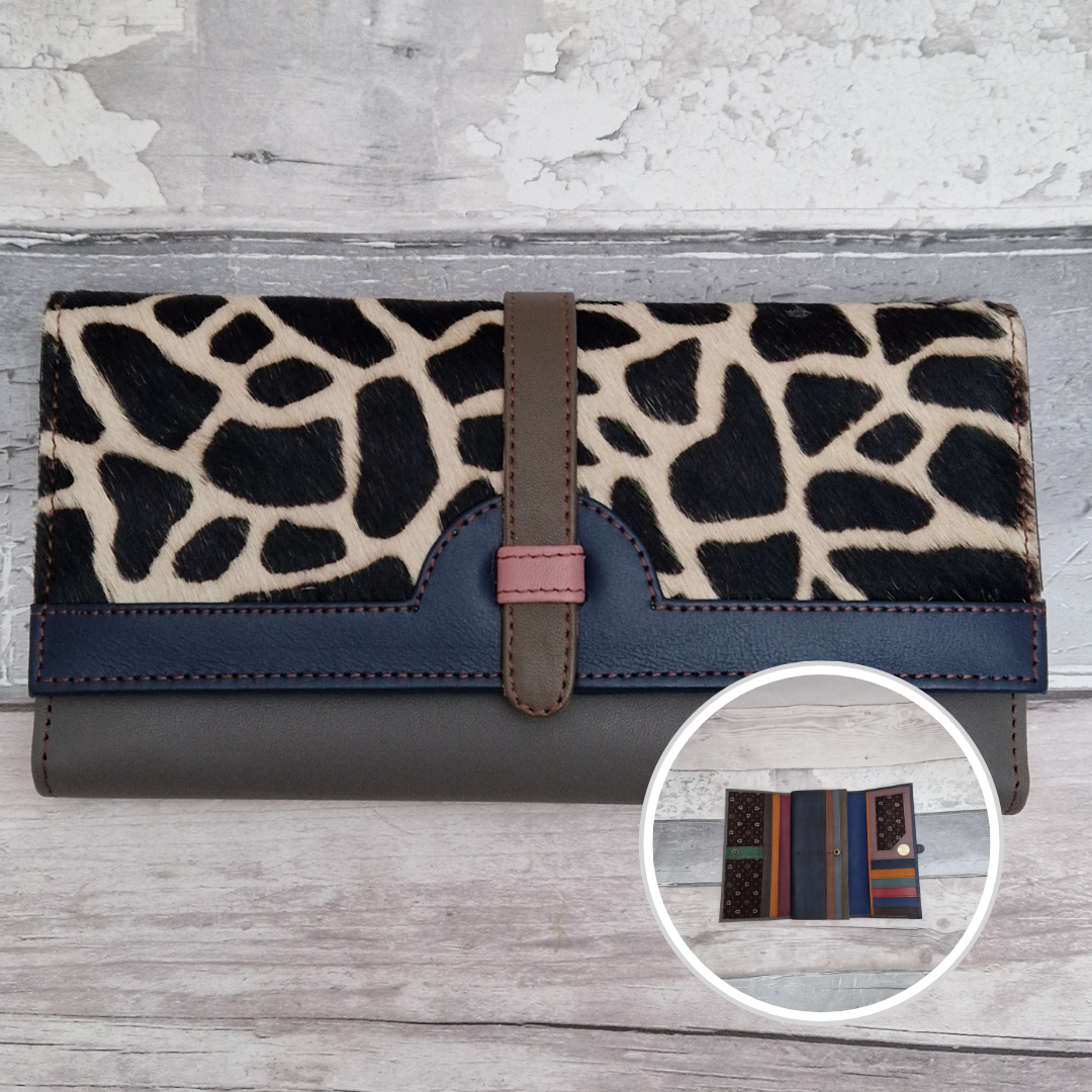 A long wallet with a giraffe-print top panel, dark navy blue band, and dark gray base, shown closed on a light wood surface. A circular inset at bottom right shows the striped interior.