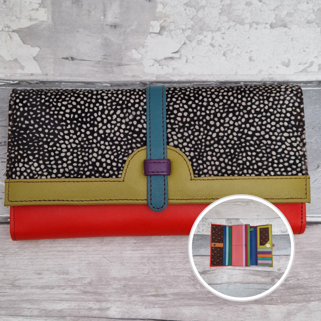 A multicolor leather wallet with a black and white spotted pattern top flap, a teal strap, a yellow-green band, and a red base, shown on a light gray wood background with a circular inset showing its interior compartments.