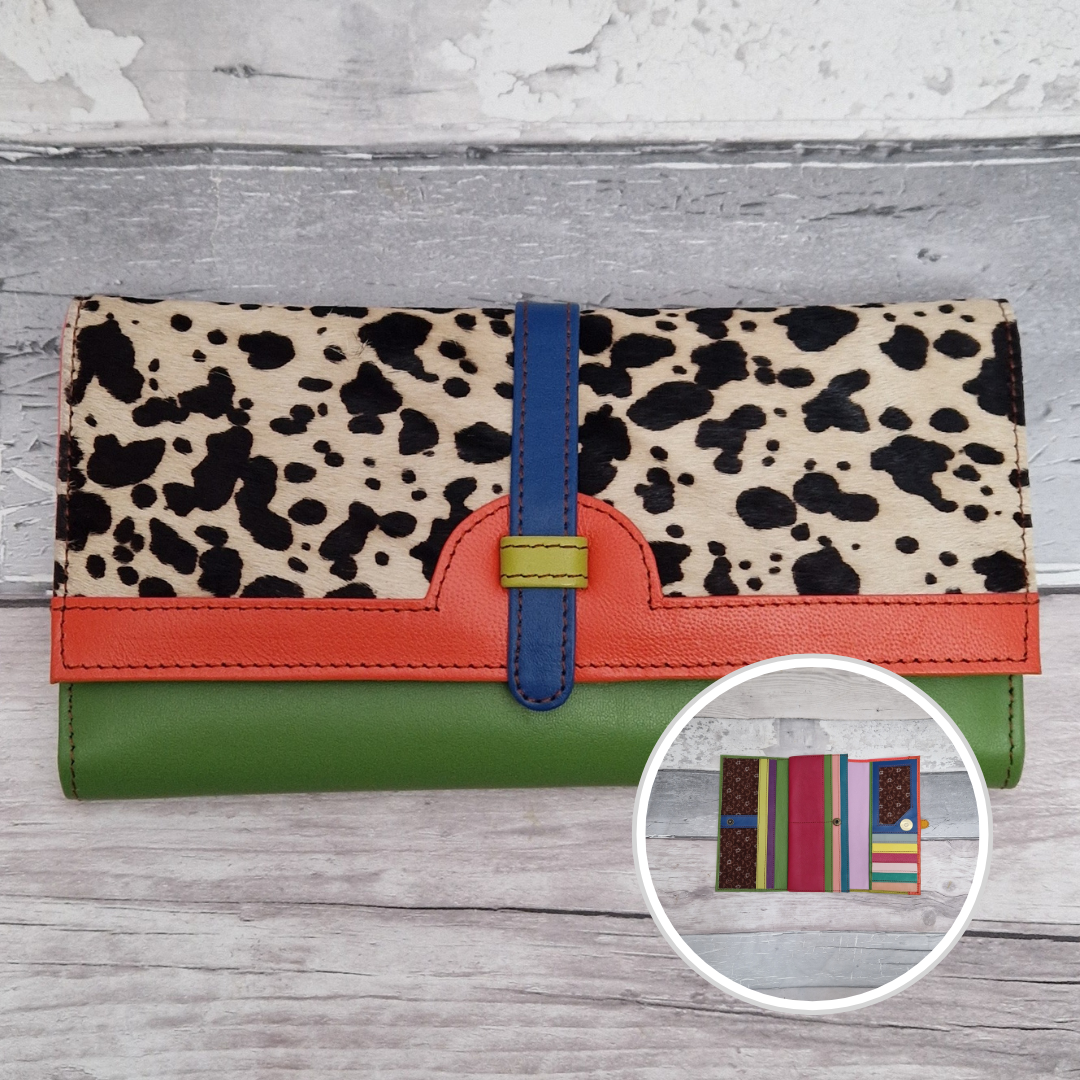 A multicolor wallet with a cow-print top half, a bright orange-red leather band, a blue strap closure, and a green base, displayed on a gray wood background with a circular inset showing fabric options.