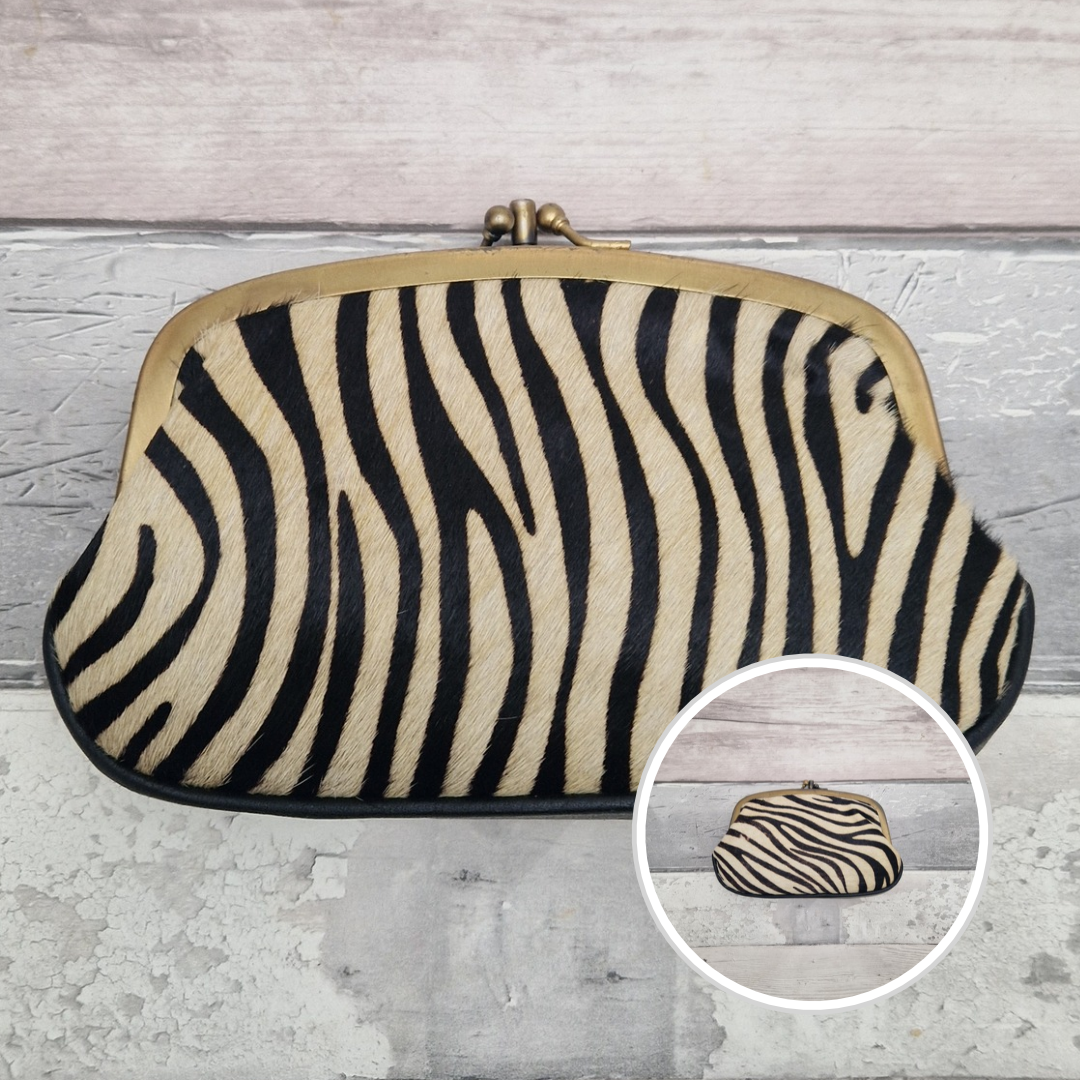 A zebra-print kissel clutch purse with a gold-tone metal frame and clasp, displayed on a light gray wood plank background with a circular inset showing the clutch closed.