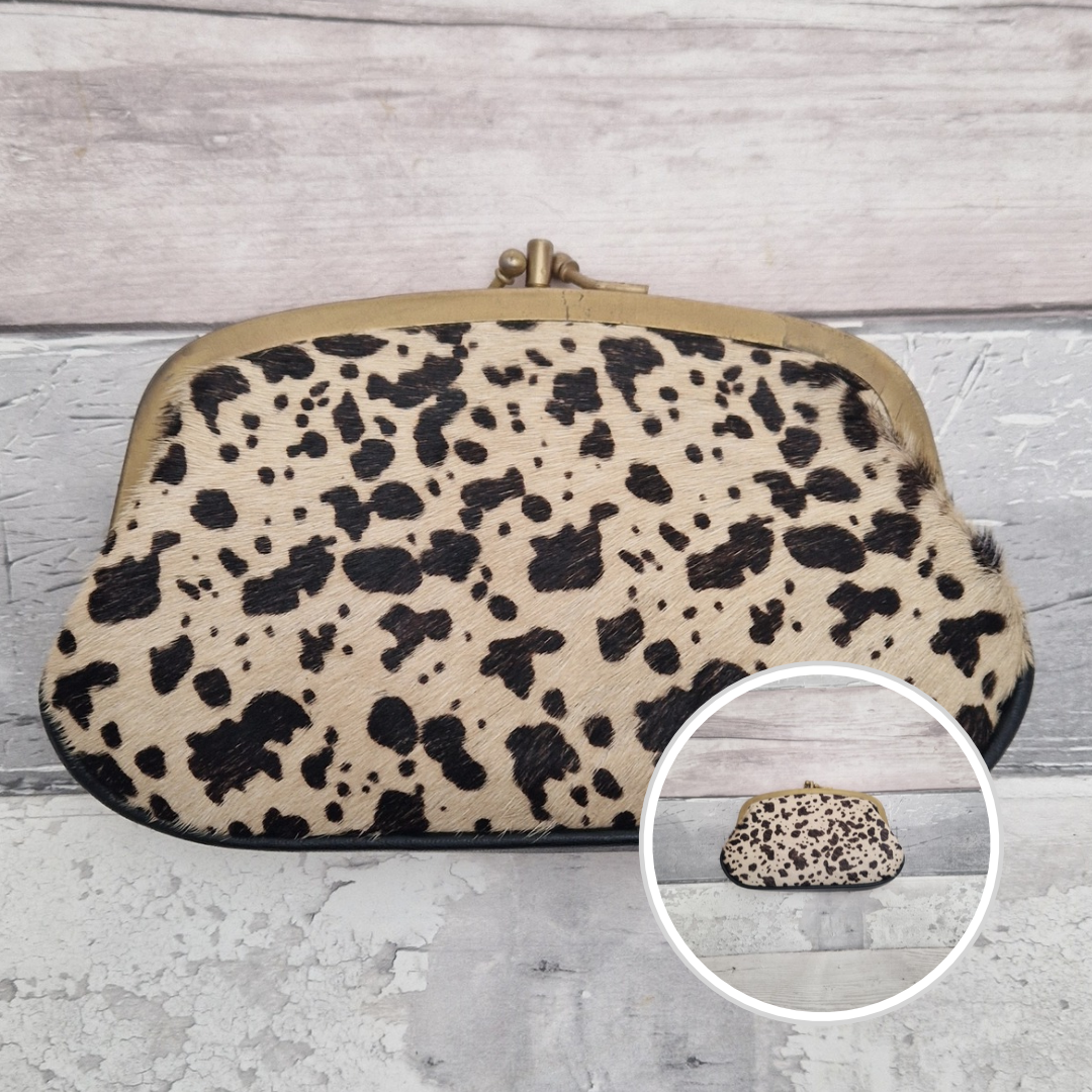 A black & white cow print hair on clutch purse with a gold-tone metal frame closure, shown on a light gray wood-plank background with a circular inset showing the product from the front.