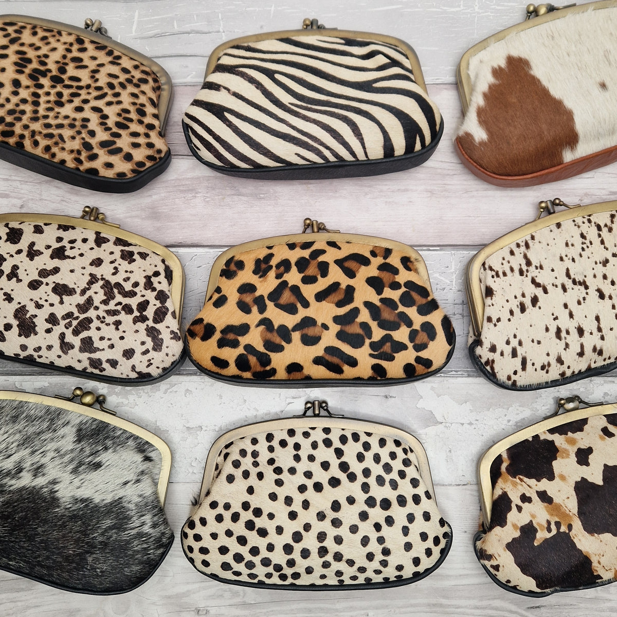 Nine small kiss-lock clutch bags arranged in a 3x3 grid on a light whitewashed wood background, each showing a different animal print pattern including leopard, zebra, cowhide, and spotted designs.