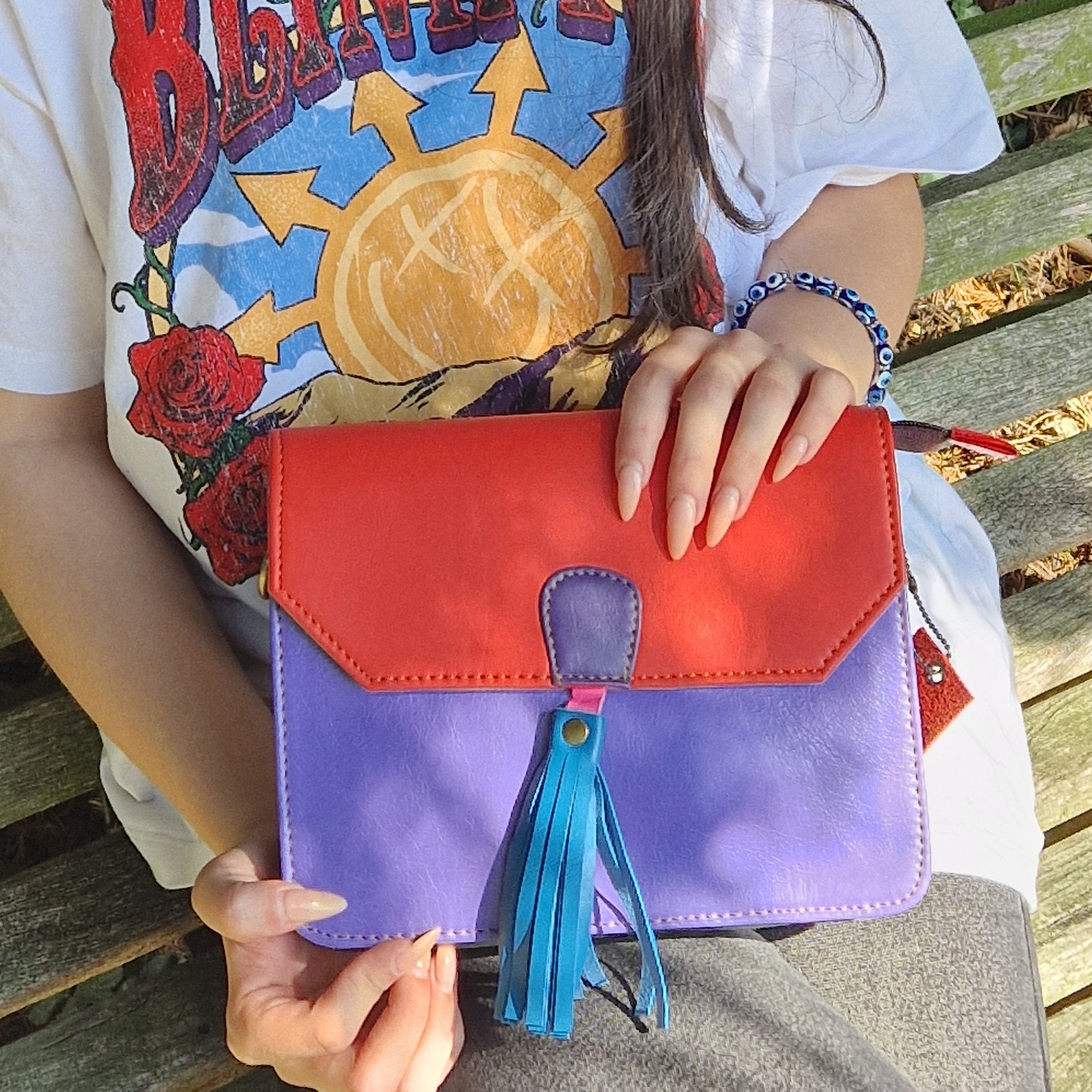 Person holding a red and purple handbag with a blue tassel on a wooden bench.