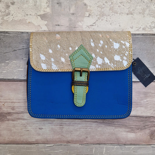 A structured handbag with a vibrant blue body and a textured beige flap speckled with white, featuring a light green leather buckle detail and brass hardware, displayed on a rustic wooden background.