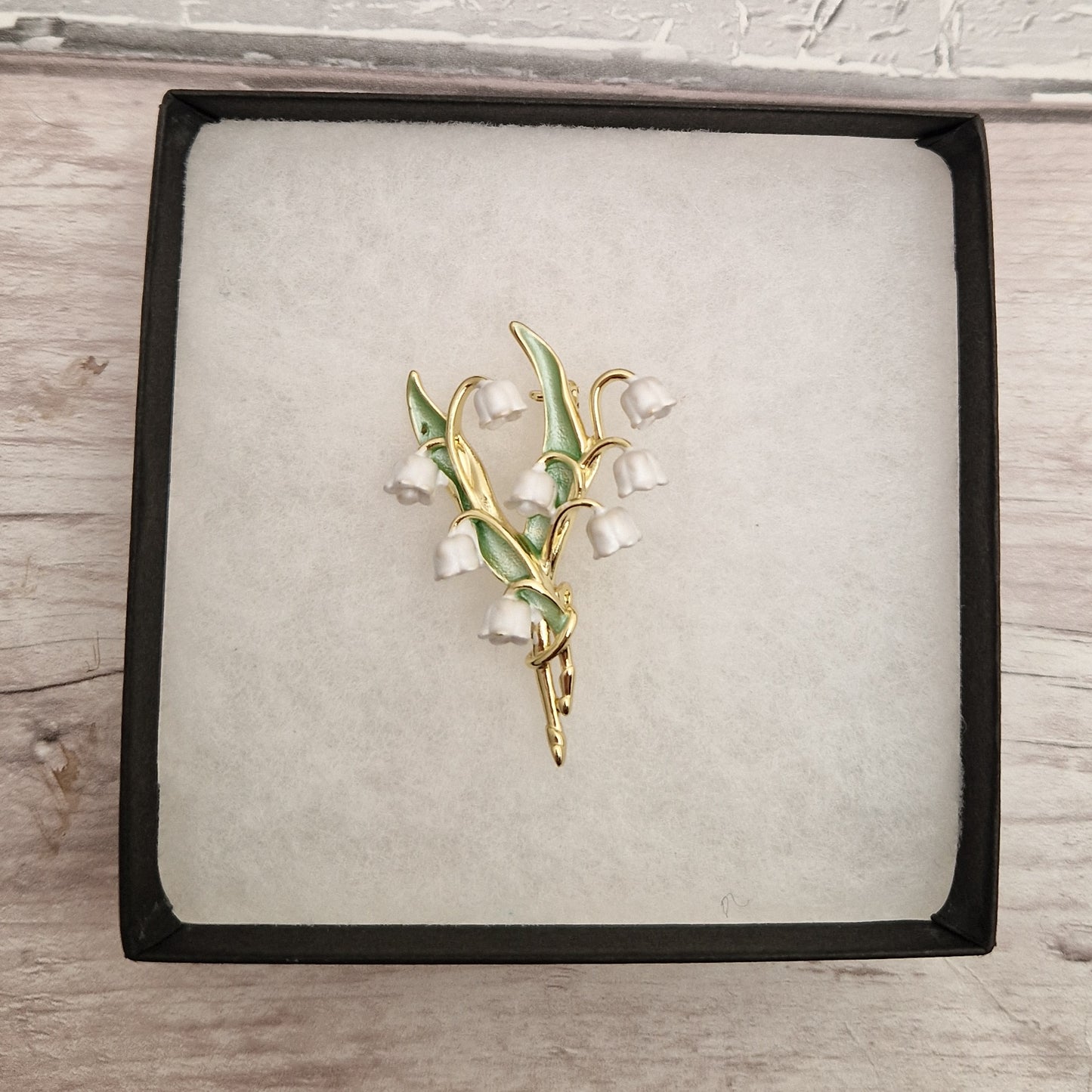 Photo of a spray of Lily of the Valley white flowers and green leaves formed as a ladies brooch with a gold stem in a gift box.