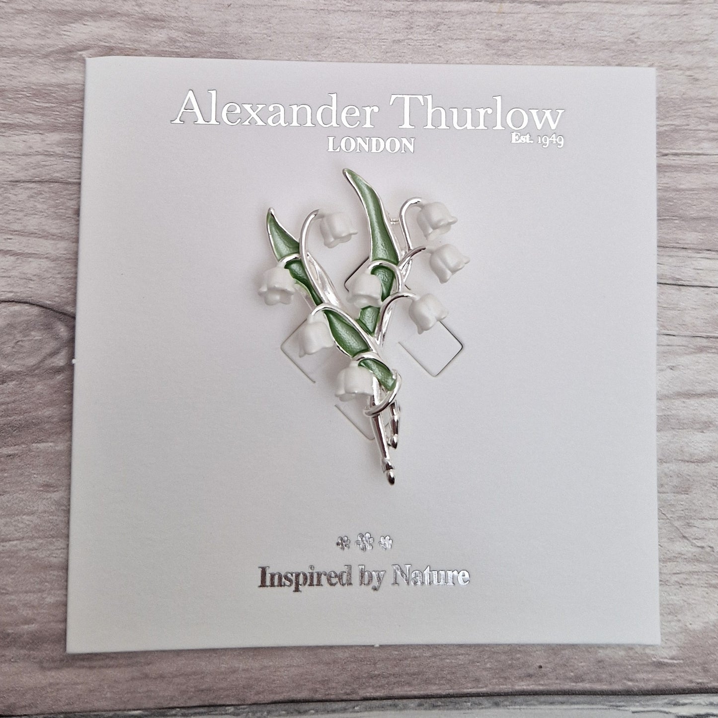 Photo of a spray of Lily of the Valley white flowers and green leaves formed as a ladies brooch with a silver stem on a display card.