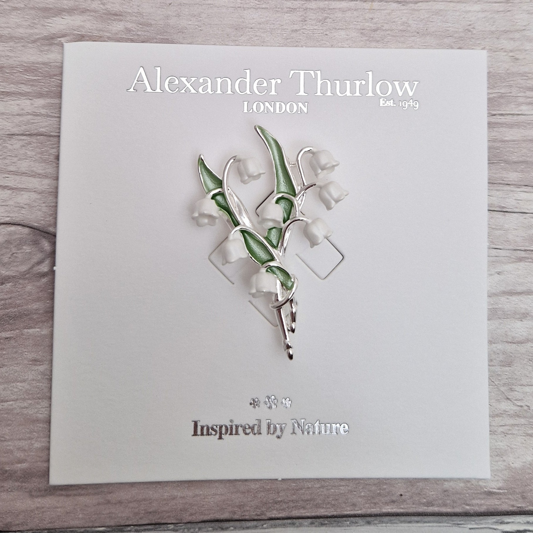 Photo of a spray of Lily of the Valley white flowers and green leaves formed as a ladies brooch with a silver stem on a display card.