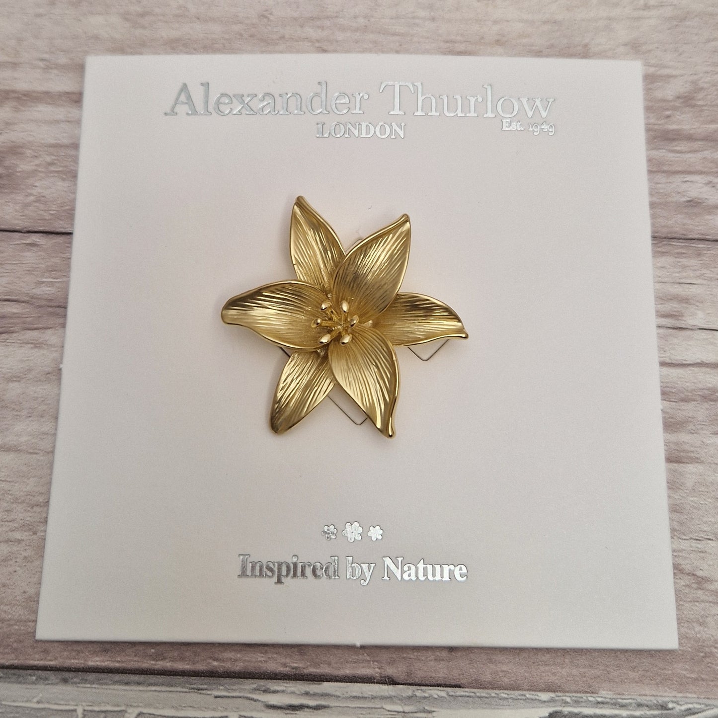 Gold flower-shaped brooch on a branded card with 'Alexander Thurlow' and 'Inspired by Nature' text.
