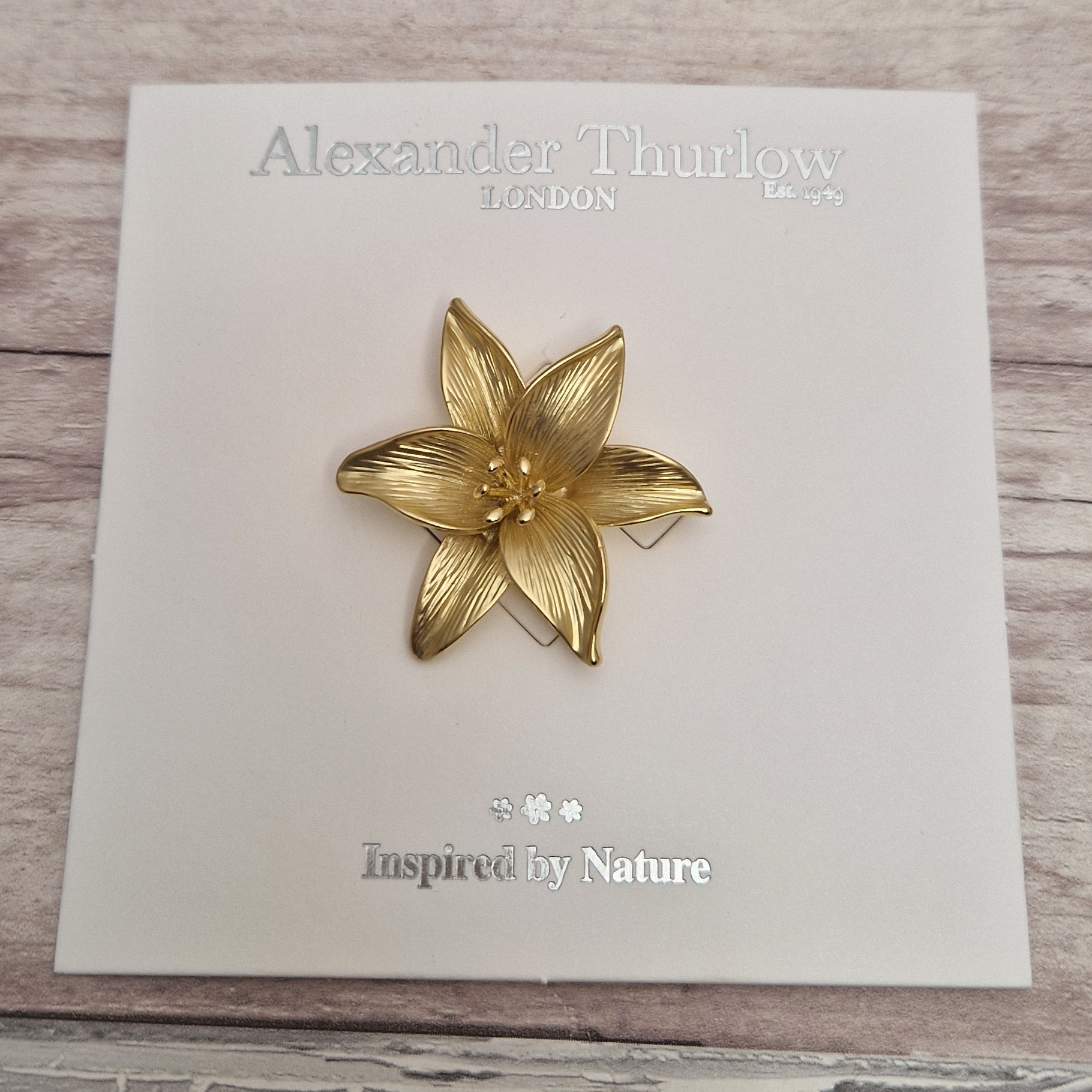 Gold flower-shaped brooch on a branded card with 'Alexander Thurlow' and 'Inspired by Nature' text.