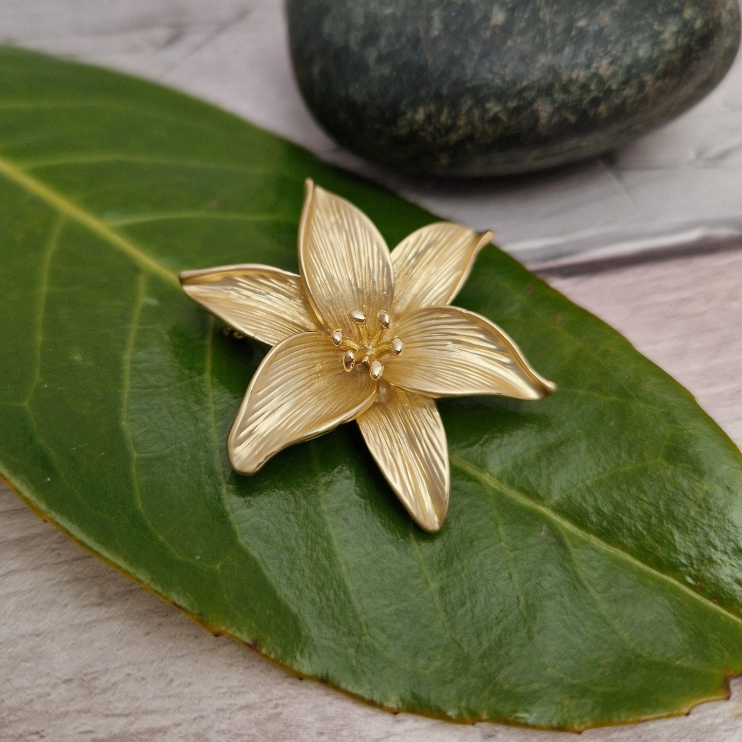 Gold Lily brooch on a green leaf with a stone in the background.