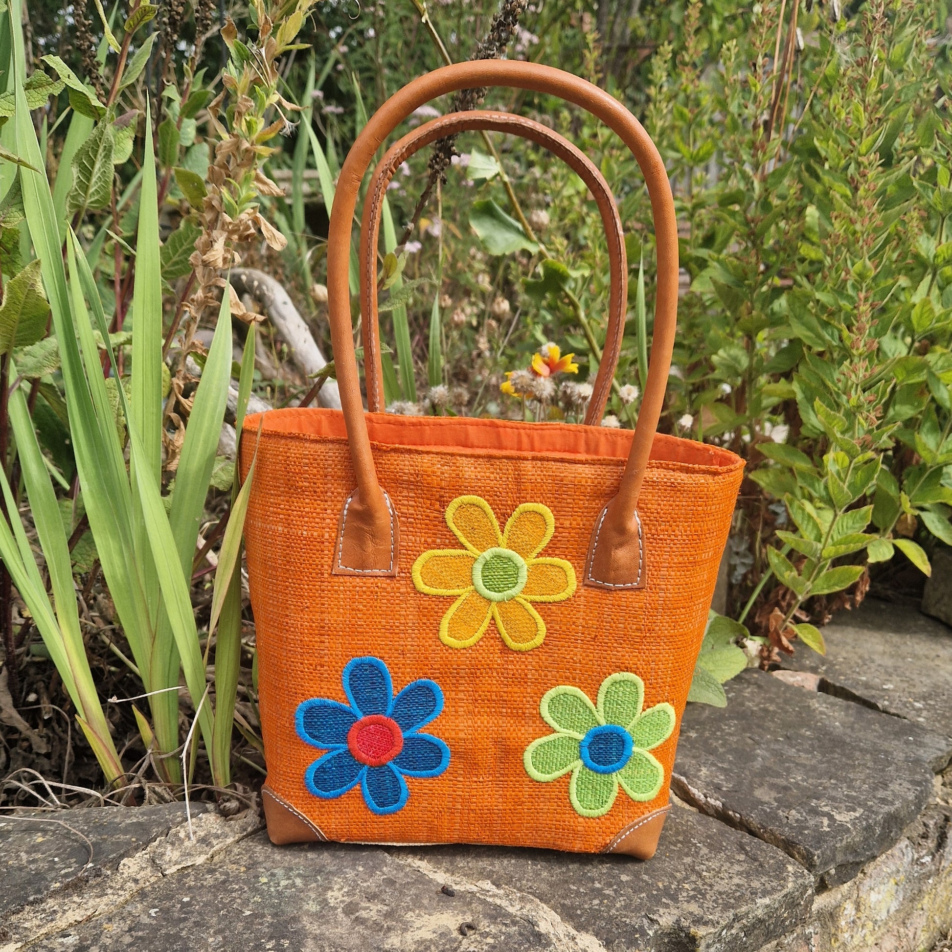 Orange handbag with flower appliqués on a stone surface with greenery in the background