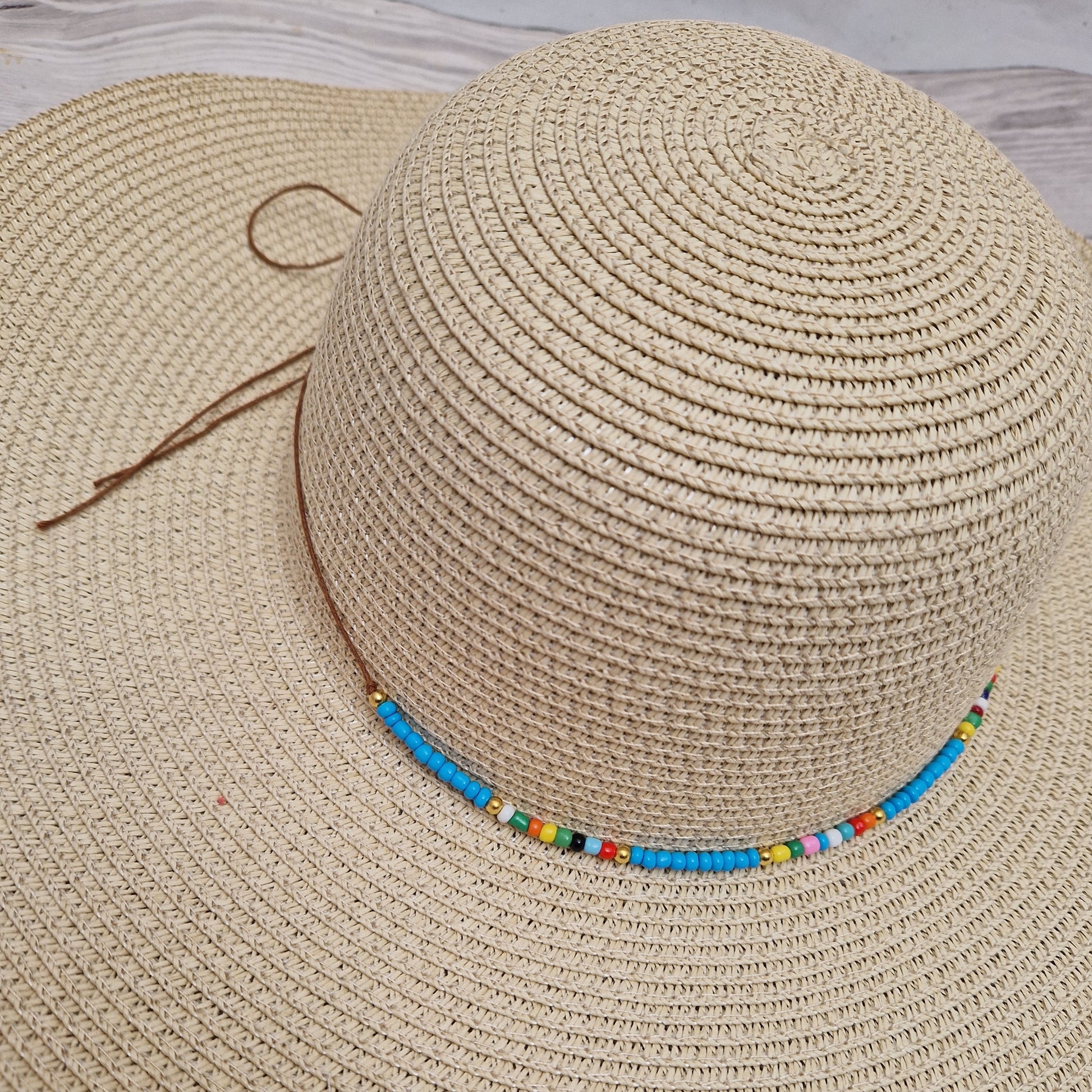 Close up view of a natural coloured wide brimmed hat with a cord around the crown covered in predominantly blue beads. 