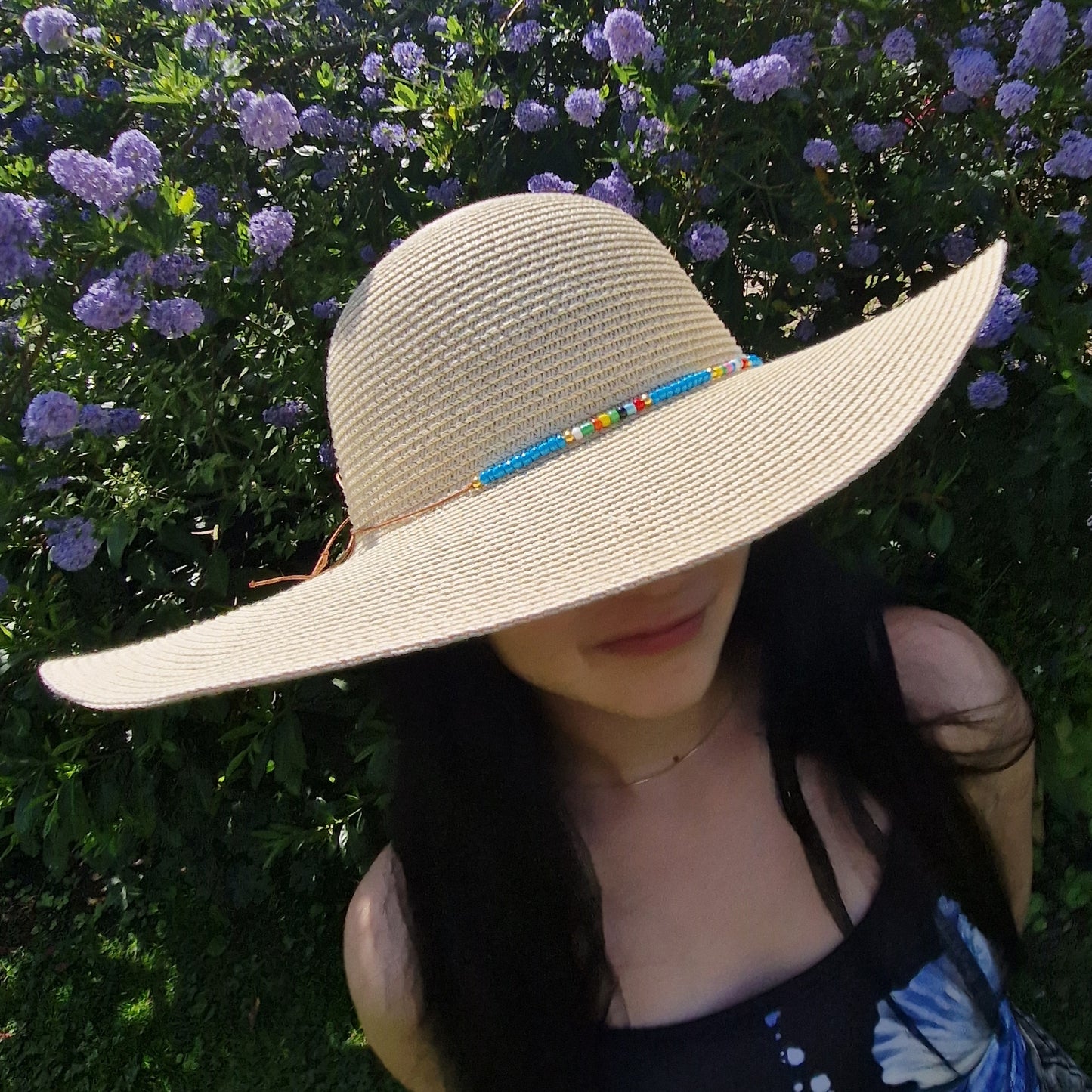 Woman wearing a natural coloured wide brimmed hat with a cord around the crown covered in predominantly blue beads. 