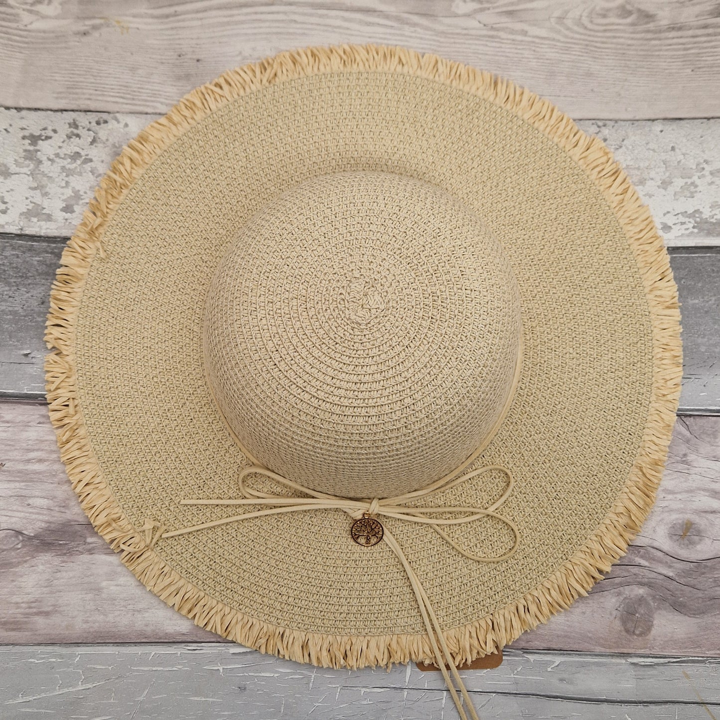 Top down view of a natural coloured wide brimmed hat with a cord around the crown tied at the back in a bow. A gold coloured tree of life charm hangs from the bow.