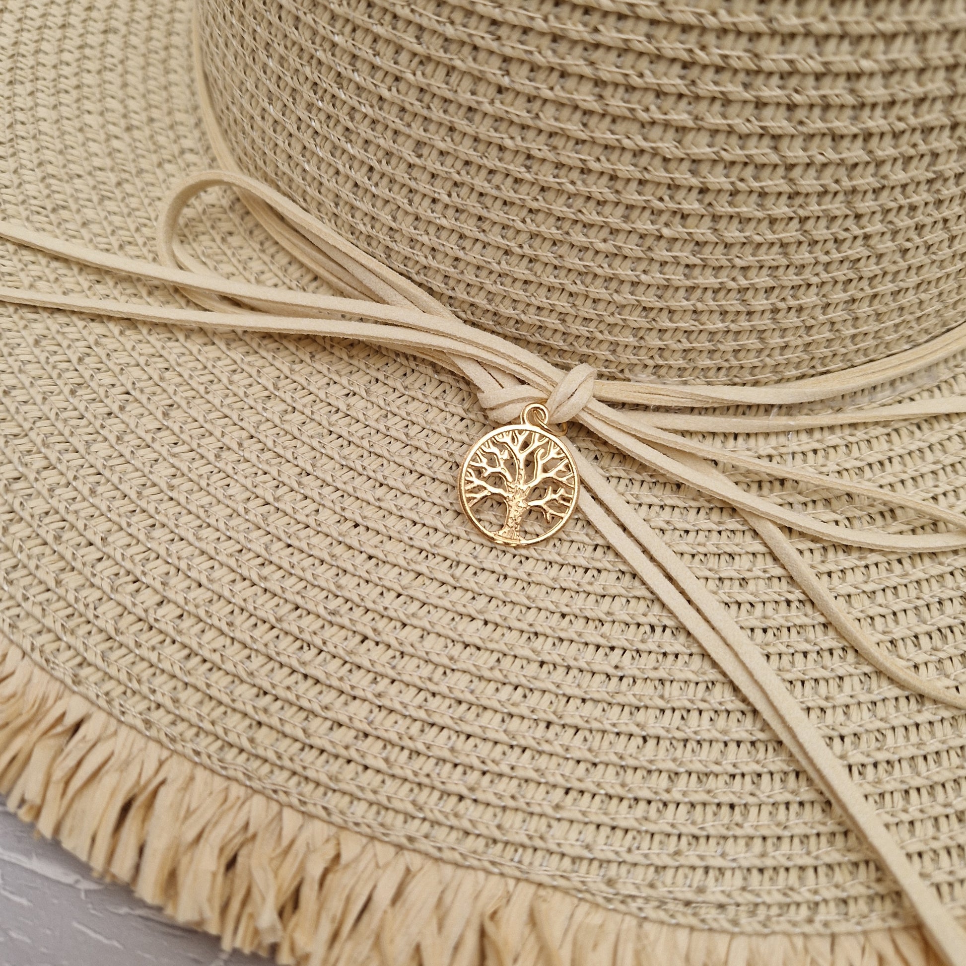 Close up view of a natural coloured wide brimmed hat with a cord around the crown tied at the back in a bow. A gold coloured tree of life charm hangs from the bow.