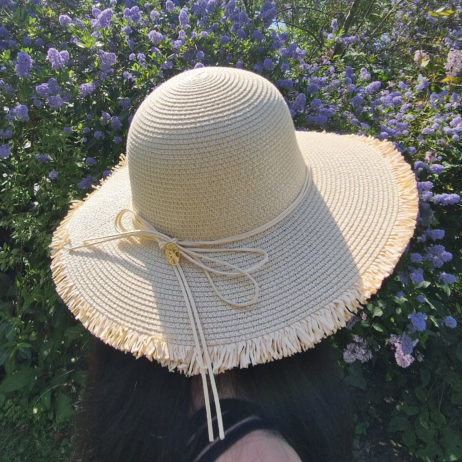 Natural coloured wide brimmed hat with a cord around the crown tied at the back in a bow. A gold coloured tree of life charm hangs from the bow.