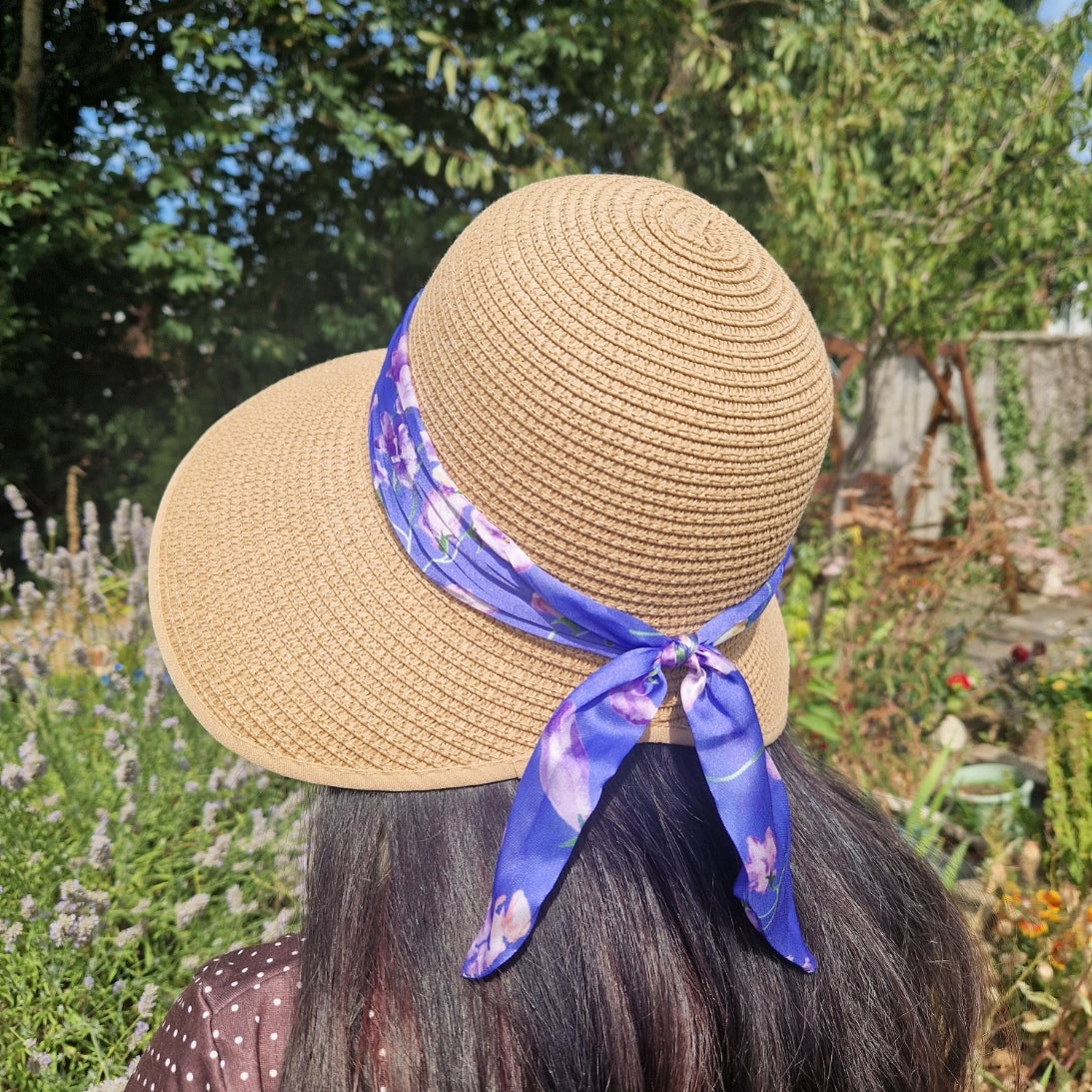 A rear view of a woman wearing a beige coloured backless bonnet with a purple floral ribbon band, photographed outdoors in a garden setting with lavender flowers and green foliage in the background.