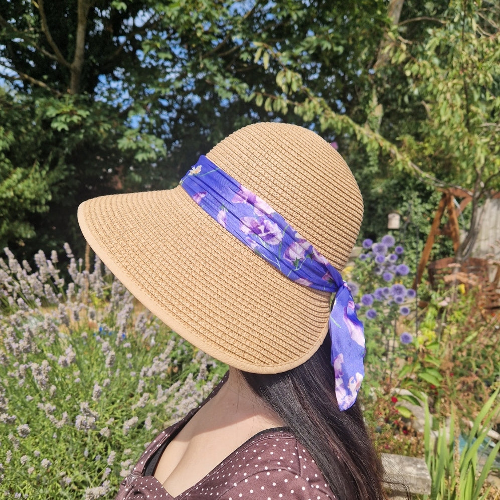 A  side view of a woman wearing a beige coloured backless bonnet with a purple floral ribbon band, photographed outdoors in a garden setting with lavender flowers and green foliage in the background.