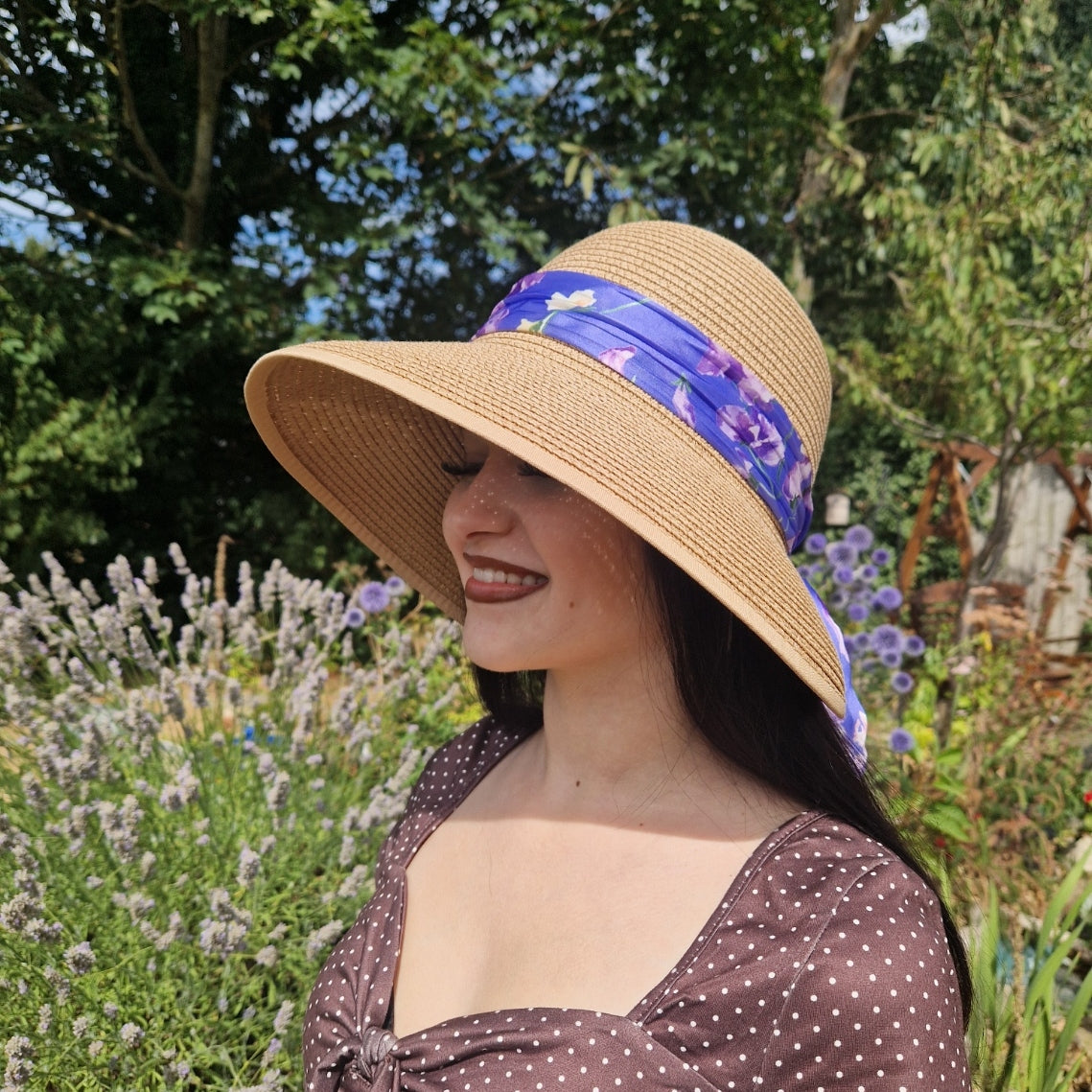 A woman wearing a beige coloured backless bonnet with a purple floral ribbon band, photographed outdoors in a garden setting with lavender flowers and green foliage in the background.