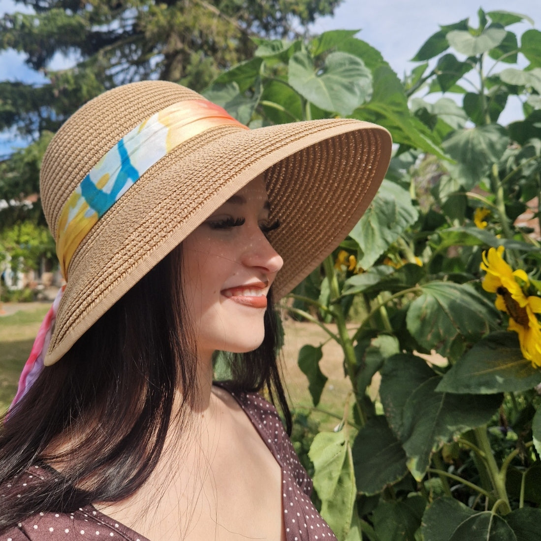 A woman wearing a beige coloured backless bonnet with a rainbow coloured ribbon band, photographed outdoors in a garden setting with sunflowers flowers and green foliage in the background.
