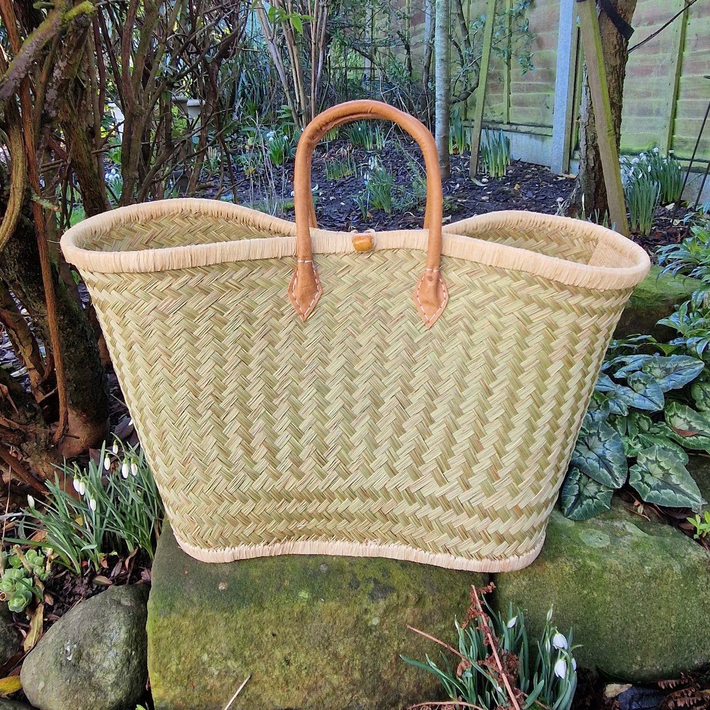 Woven basket with leather handles on a stone surface in a garden setting