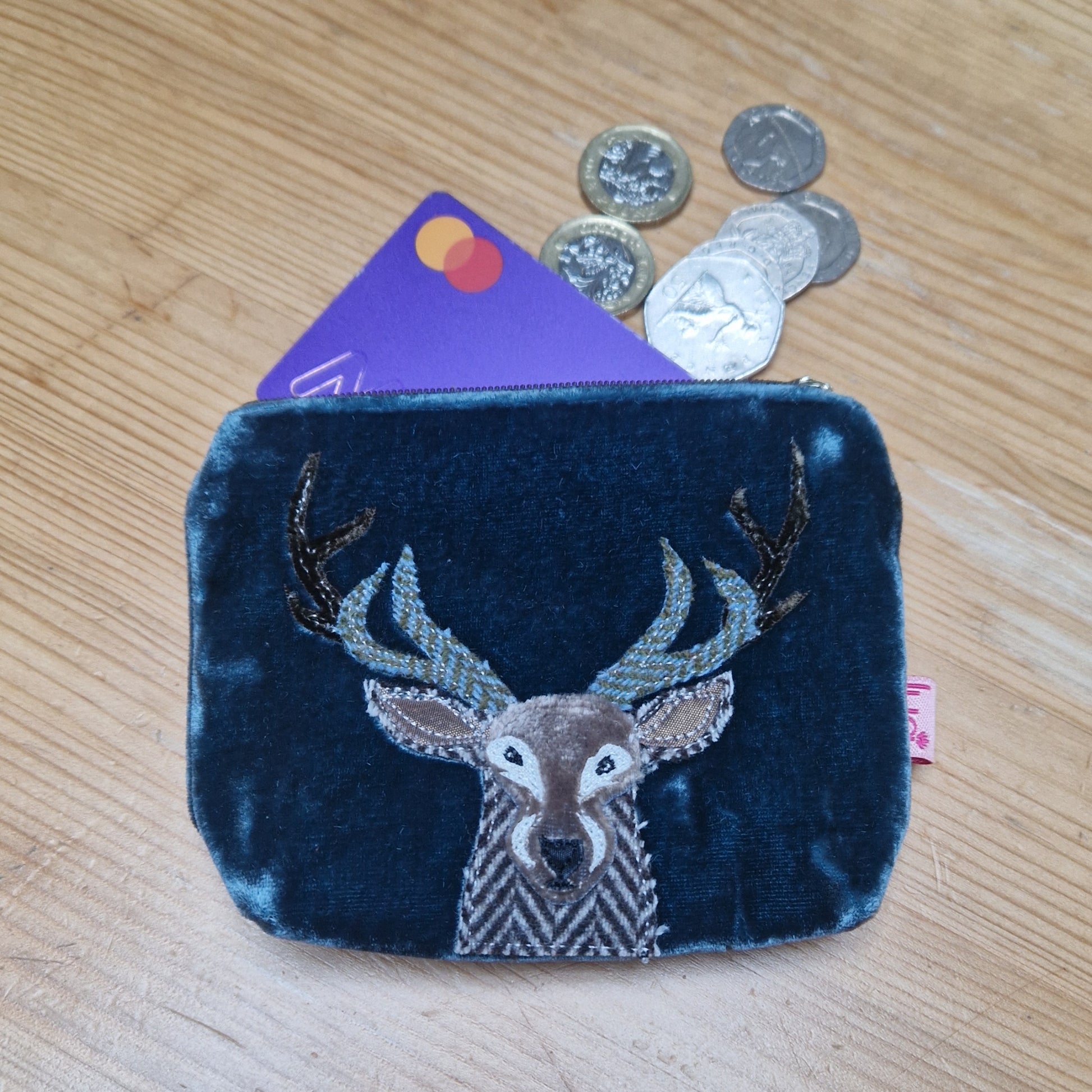 Blue velvet coin purse with deer design on a wooden surface with coins and a card.