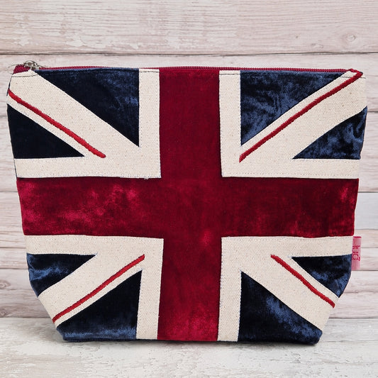 A zippered cosmetic pouch featuring a Union Jack flag design in red, navy blue, and cream velvet fabric, set against a light wood plank background.
