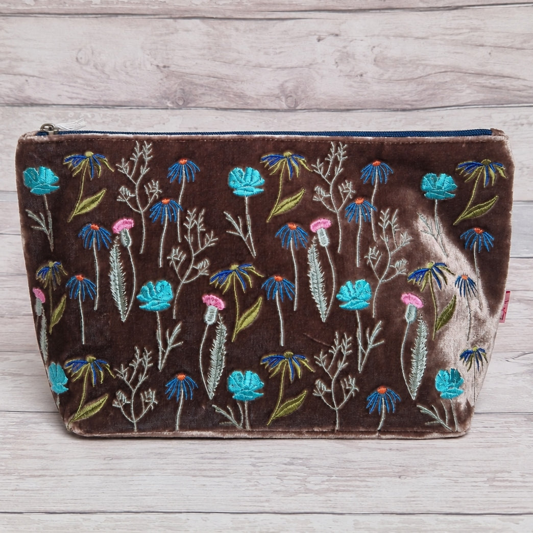 A brown velvet zippered pouch decorated with colorful embroidered flowers in blue, pink, and green, displayed on a light gray wood plank background.