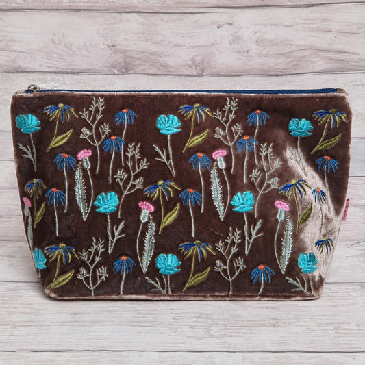 A brown velvet zippered pouch decorated with colorful embroidered flowers in blue, pink, and green, displayed on a light gray wood plank background.