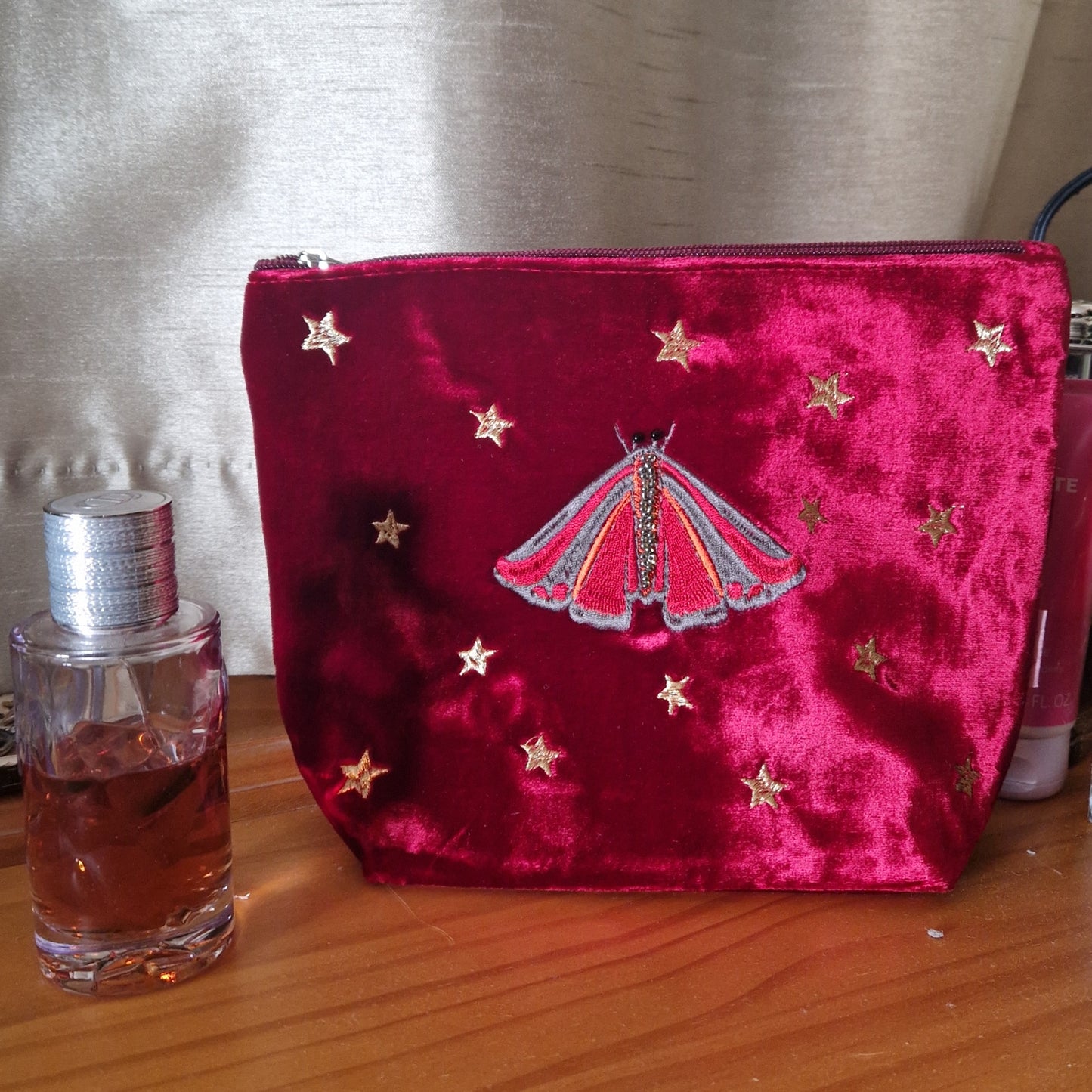 Red velvet pouch with embroidered design on a wooden surface next to a perfume bottle.