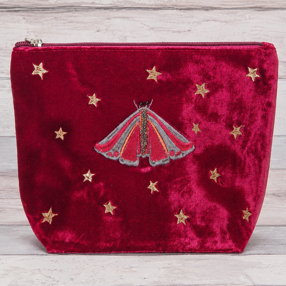 A deep red velvet zippered pouch featuring an embroidered red and gold moth in the center and scattered gold star motifs, displayed on a light gray wood background.