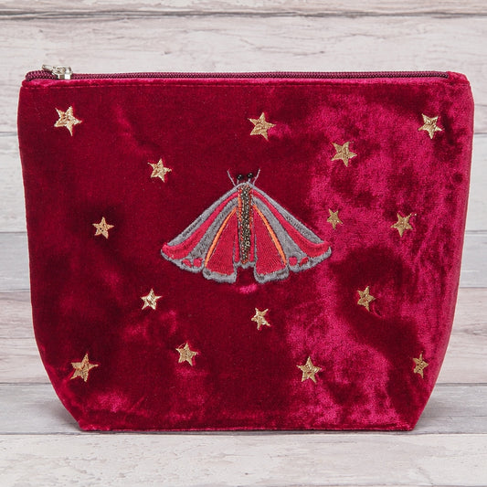 A deep red velvet zippered pouch featuring an embroidered red and gold moth in the center and scattered gold star motifs, displayed on a light gray wood background.