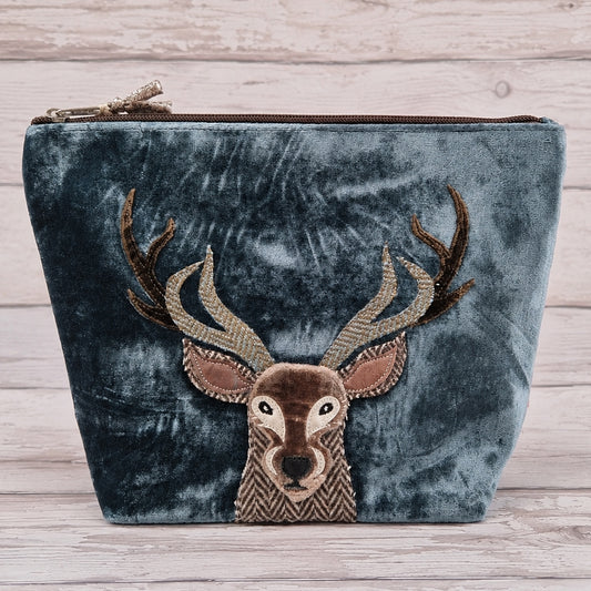 A dark teal velvet zippered pouch featuring an embroidered deer head with brown antlers on the front, set against a light wood plank background.