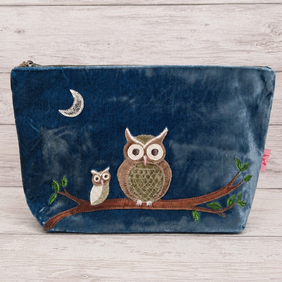 A navy blue velvet pouch featuring an embroidered design of two owls on a branch under a crescent moon, set against a light wood background.