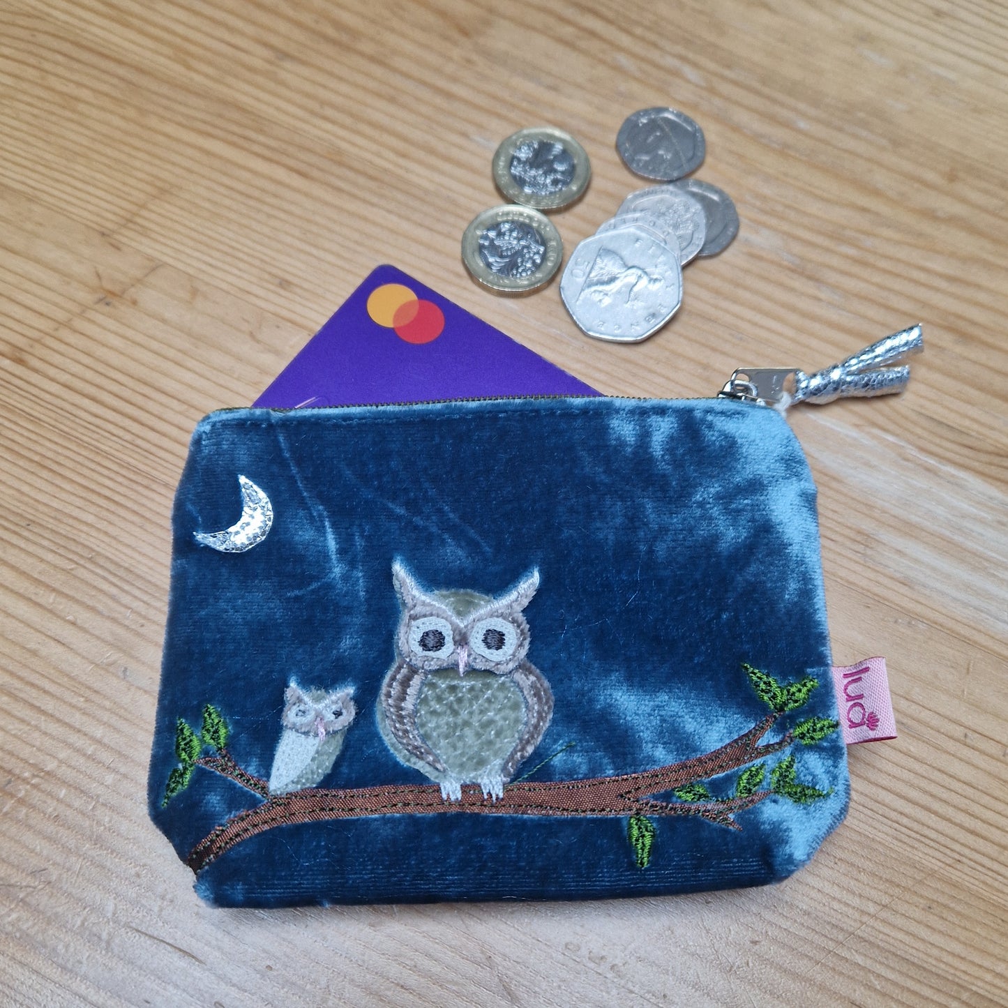 Blue coin purse with owl design on a wooden surface with coins and a card.
