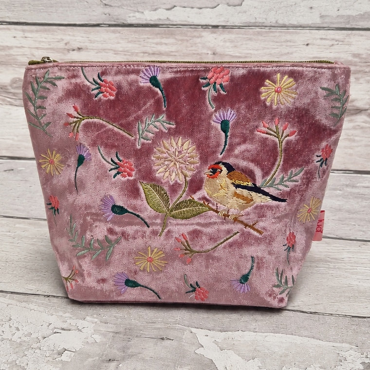A dusty rose velvet zippered pouch decorated with colorful embroidered flowers, leaves, and a small goldfinch bird, displayed on a rustic white-washed wooden surface.